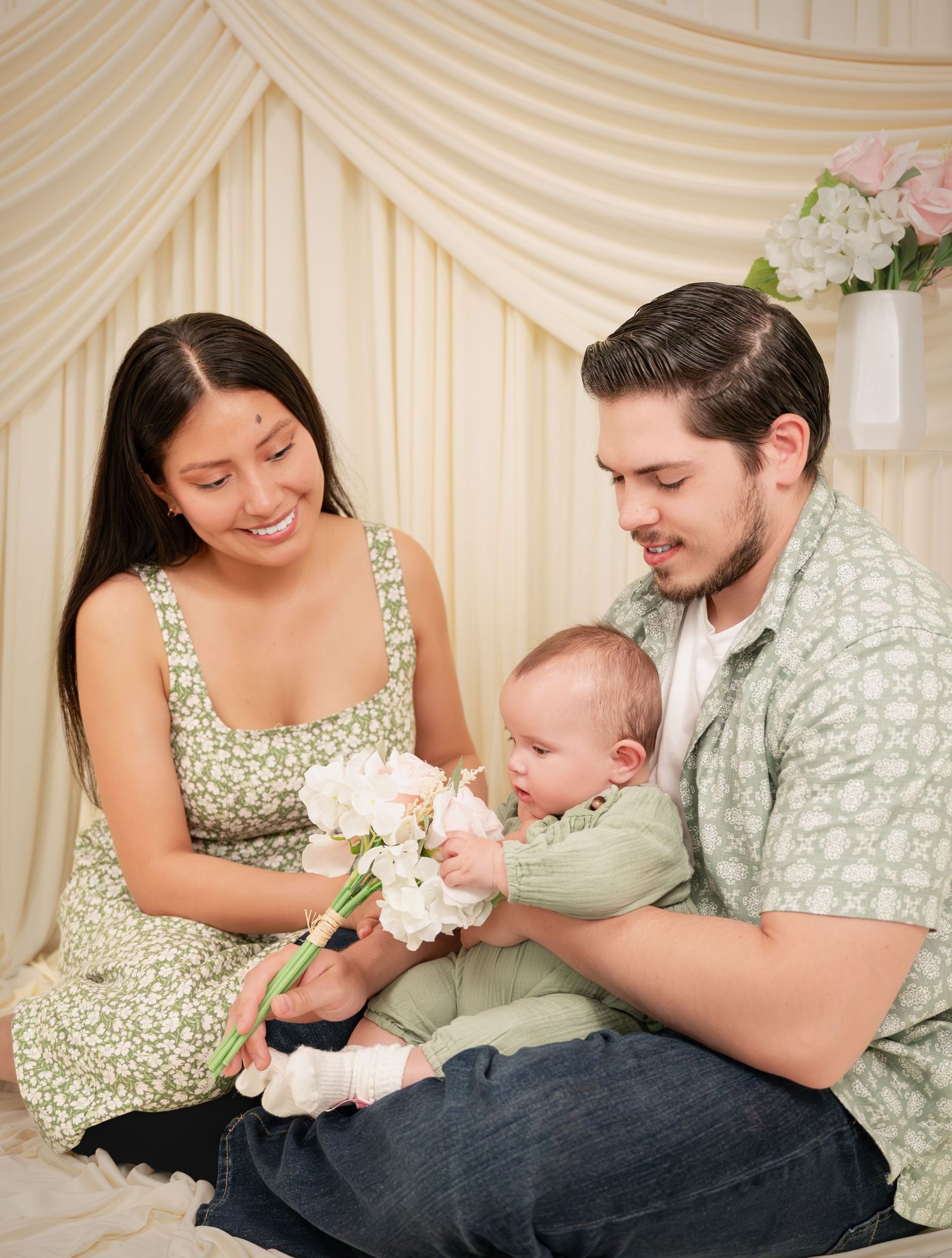 Young family with mother in green dress and father holding newborn baby with white flowers in soft studio setting.