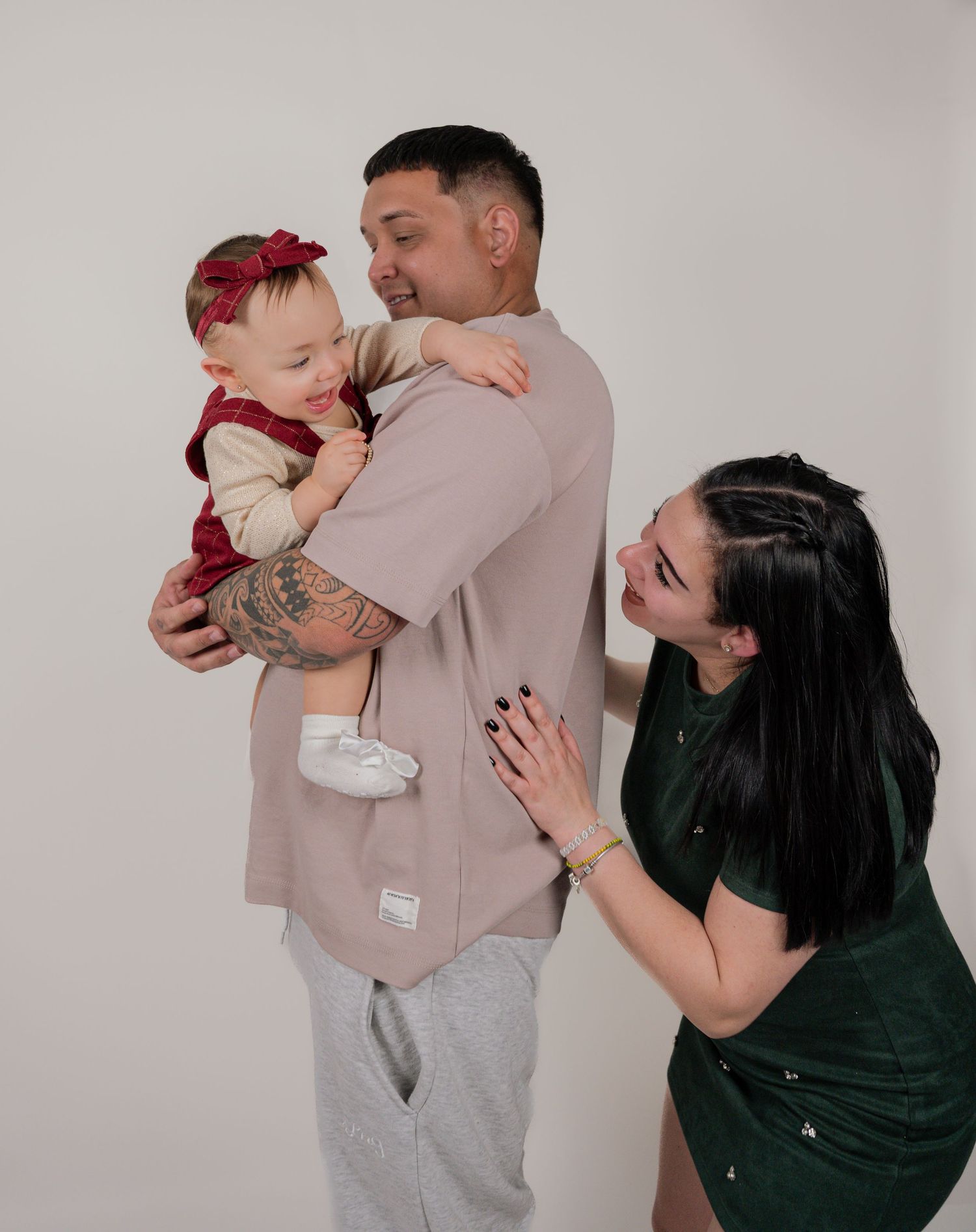 A young family poses in a studio, father holding a baby girl in a red bow while mother smiles adoringly beside them.