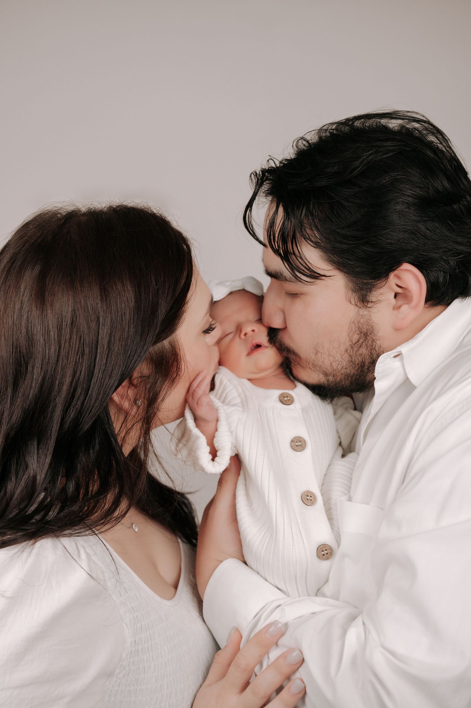 Two parents in white clothing lovingly kiss their newborn baby, captured in a tender family portrait.