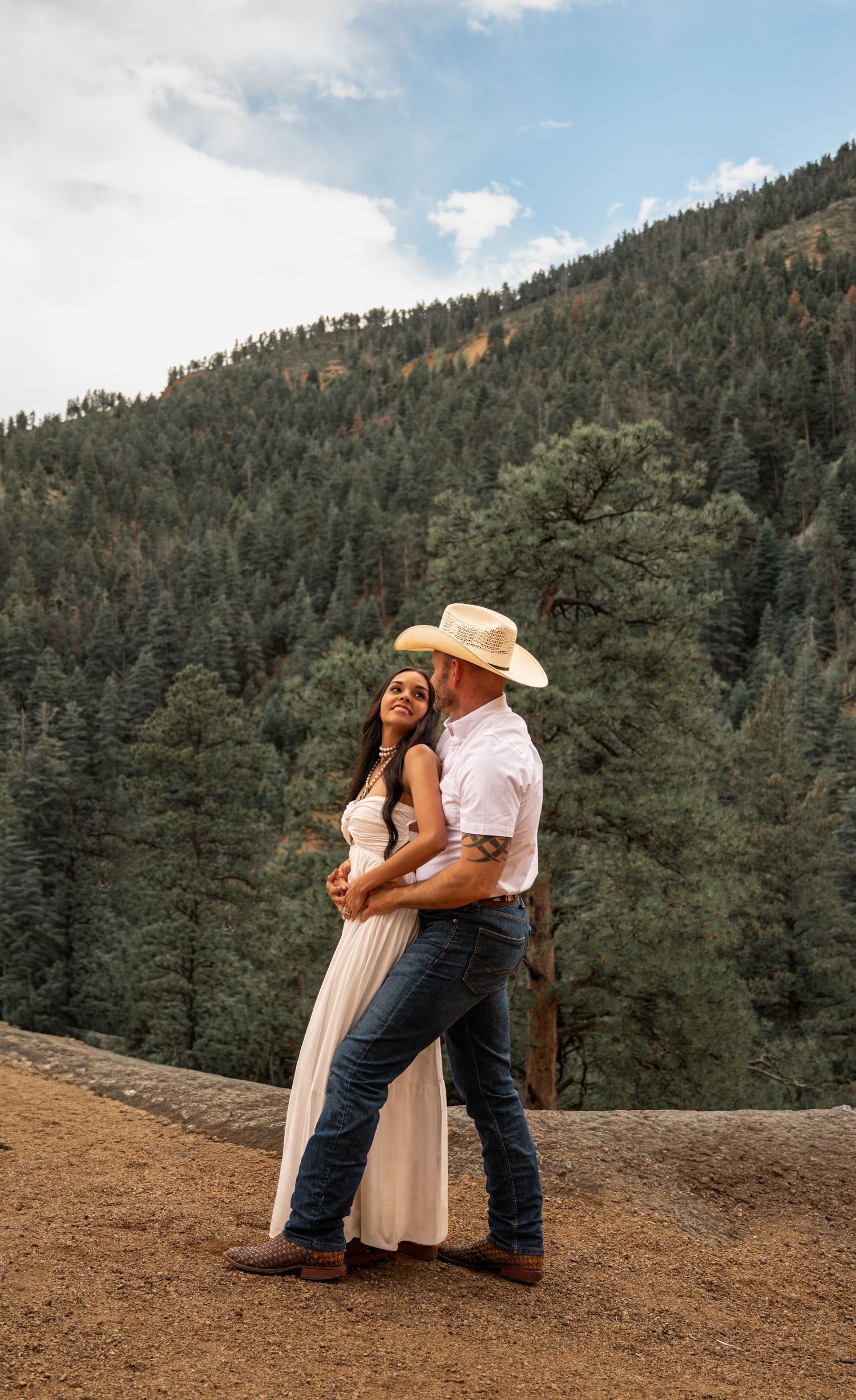 Couple embracing on a rocky overlook with lush pine forest mountains backdrop, woman in white dress, man in cowboy hat.