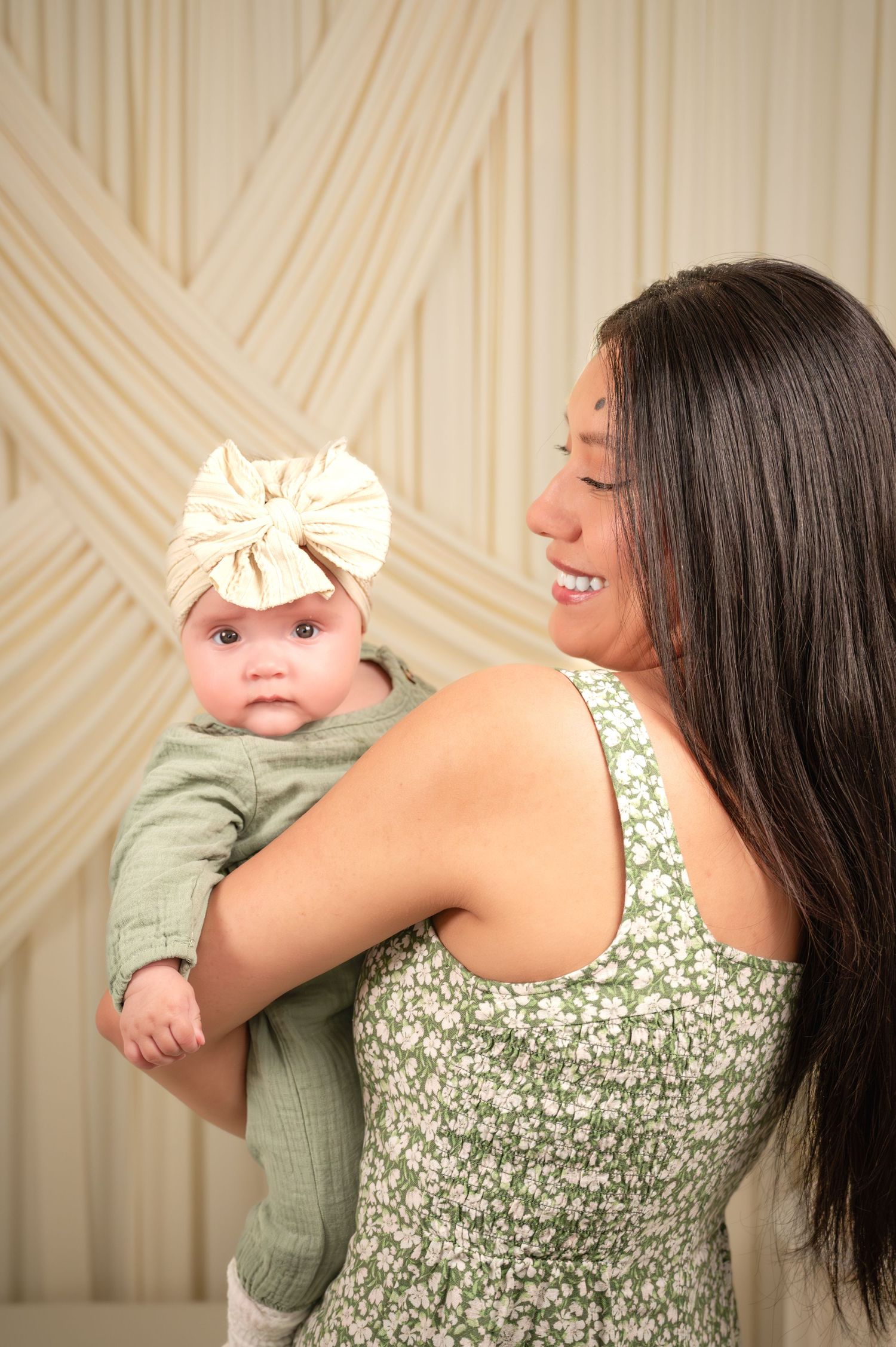 Smiling woman in green floral dress holding a baby wearing a cream bow headband against a geometric backdrop.
