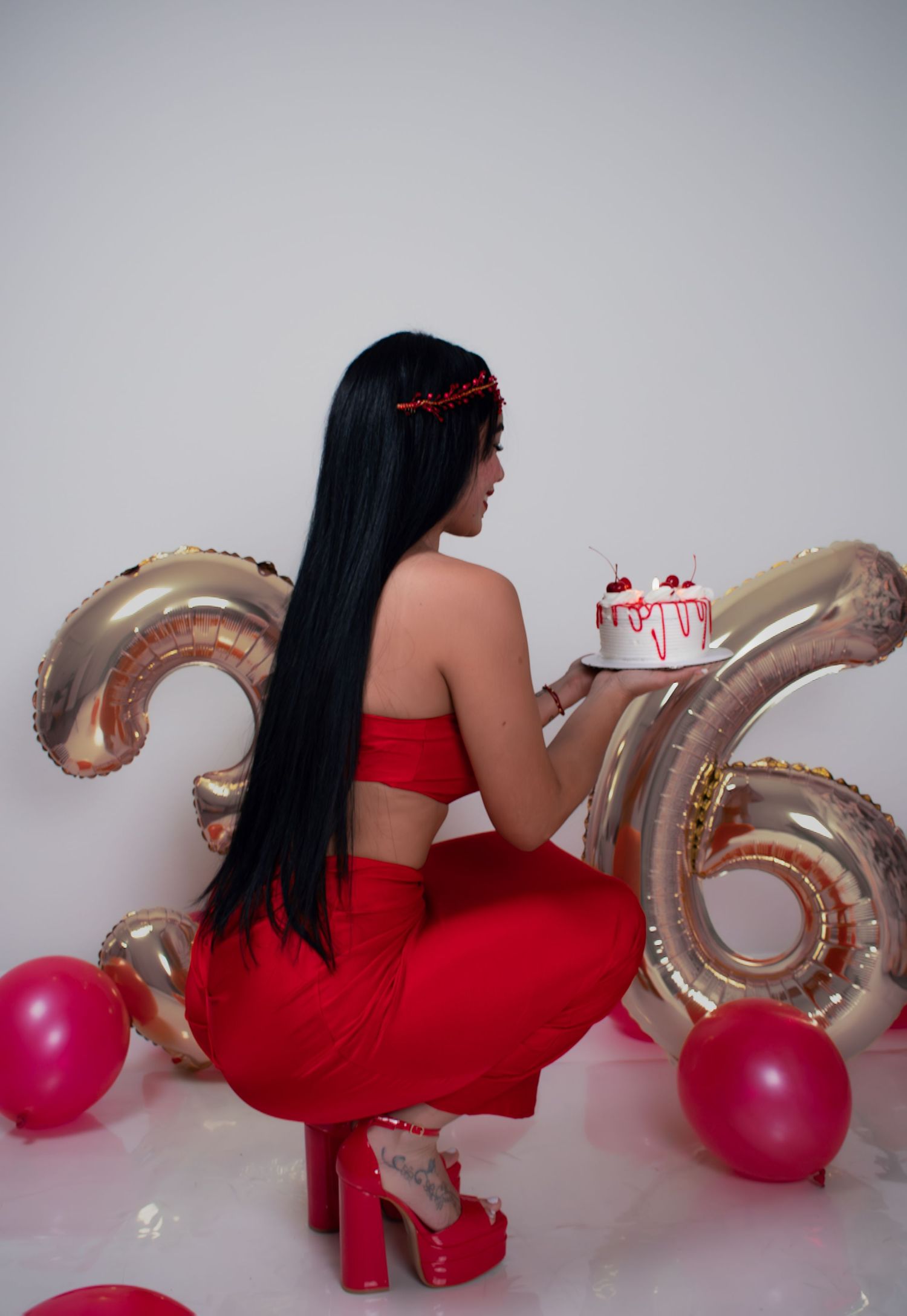 Woman in red outfit with long black hair holding birthday cake beside gold number 36 balloons.