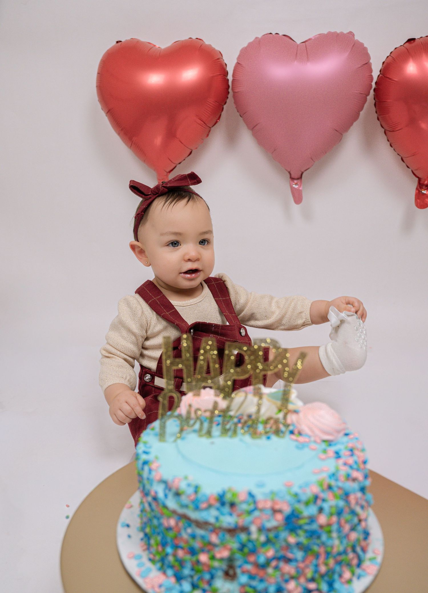 Baby girl in bow headband touches her first birthday cake with heart balloons behind her.