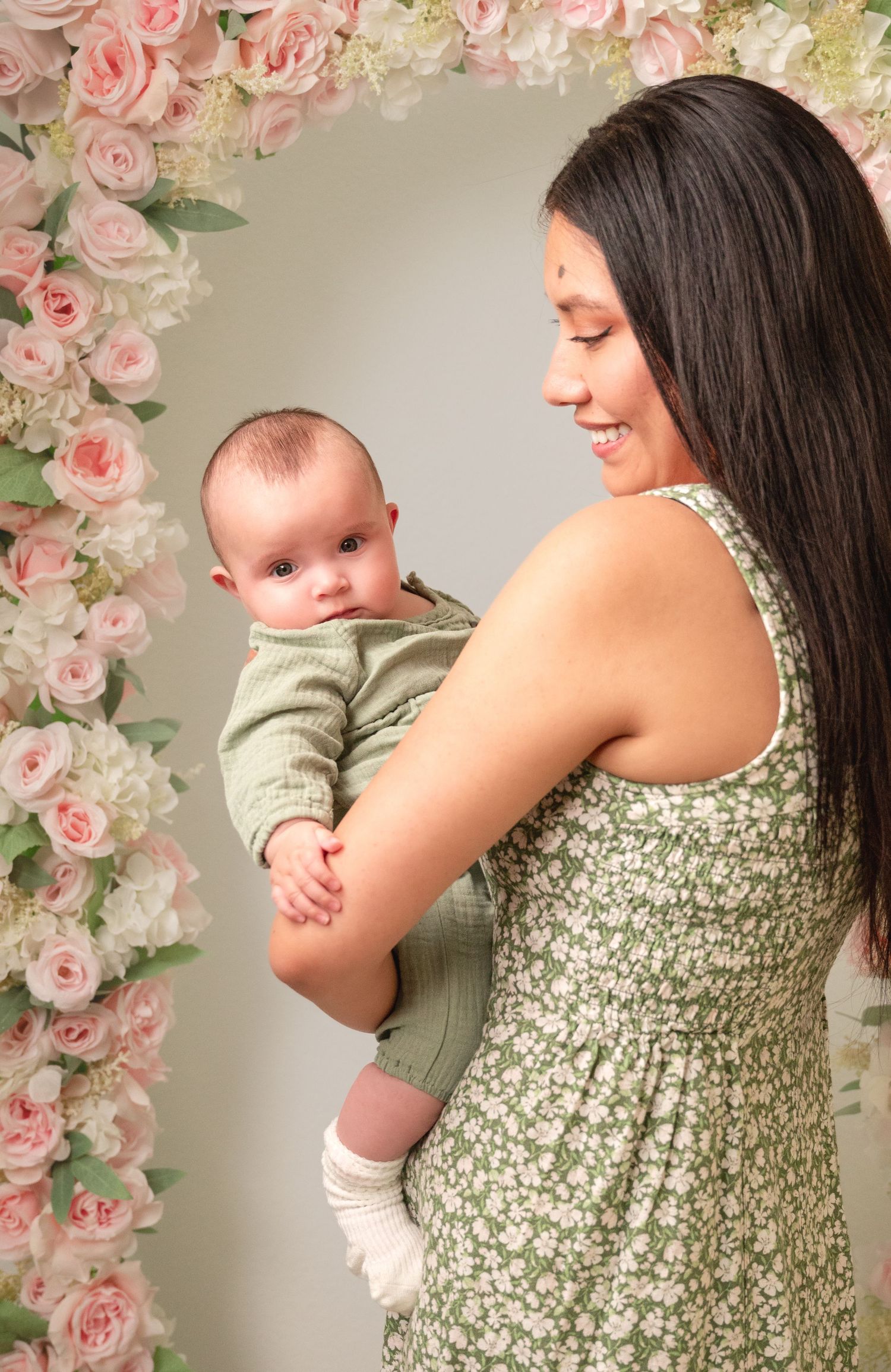 A smiling mother in a green floral dress holds her newborn baby in front of a pink rose arch backdrop.