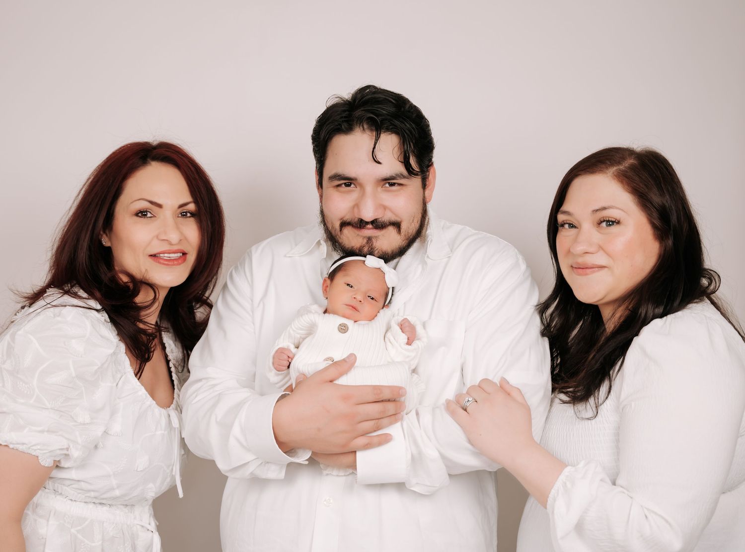 Family portrait of two women and a man holding a newborn baby, all dressed in white against a light background.