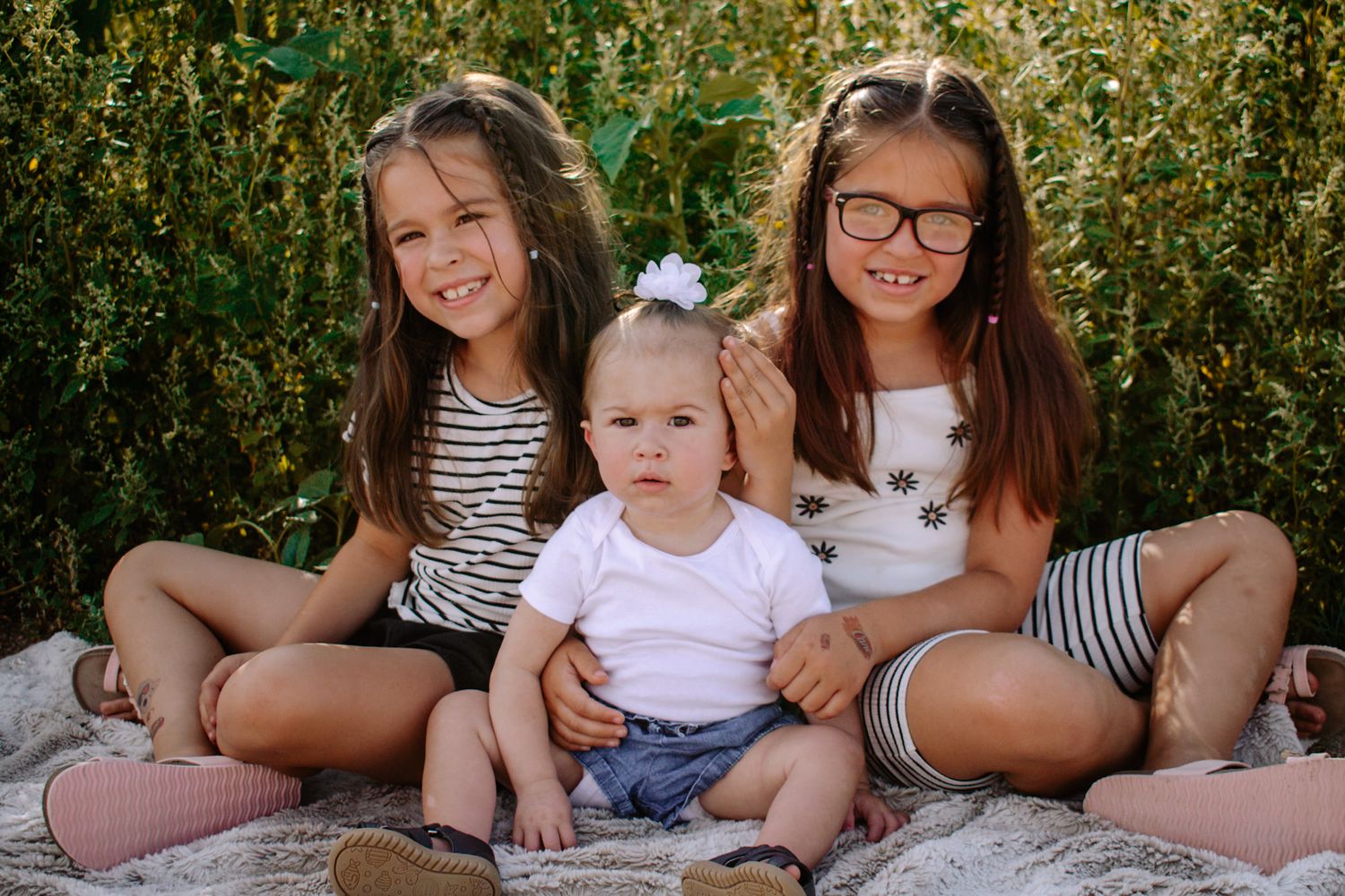 Two older sisters smile while sitting with their baby sibling on a blanket outdoors.