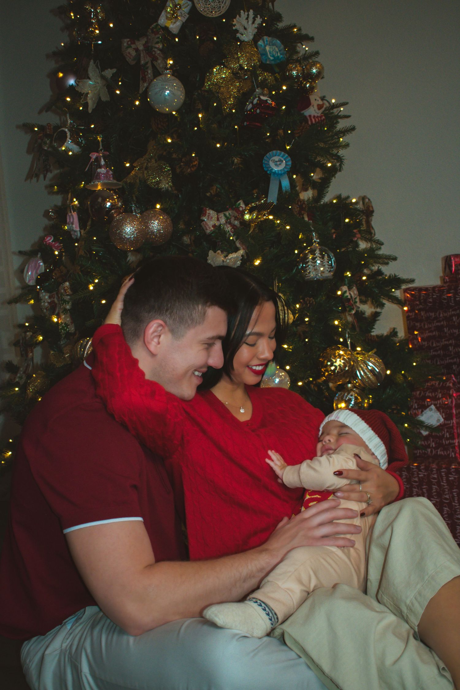 Happy couple in red outfits holding newborn baby in front of decorated Christmas tree.