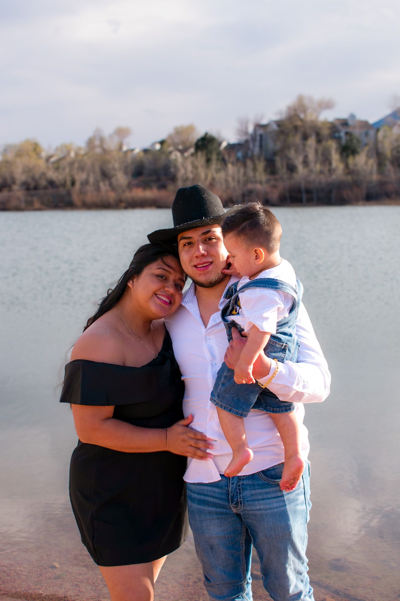 A young family of three poses by a lake, with the father holding their baby son.