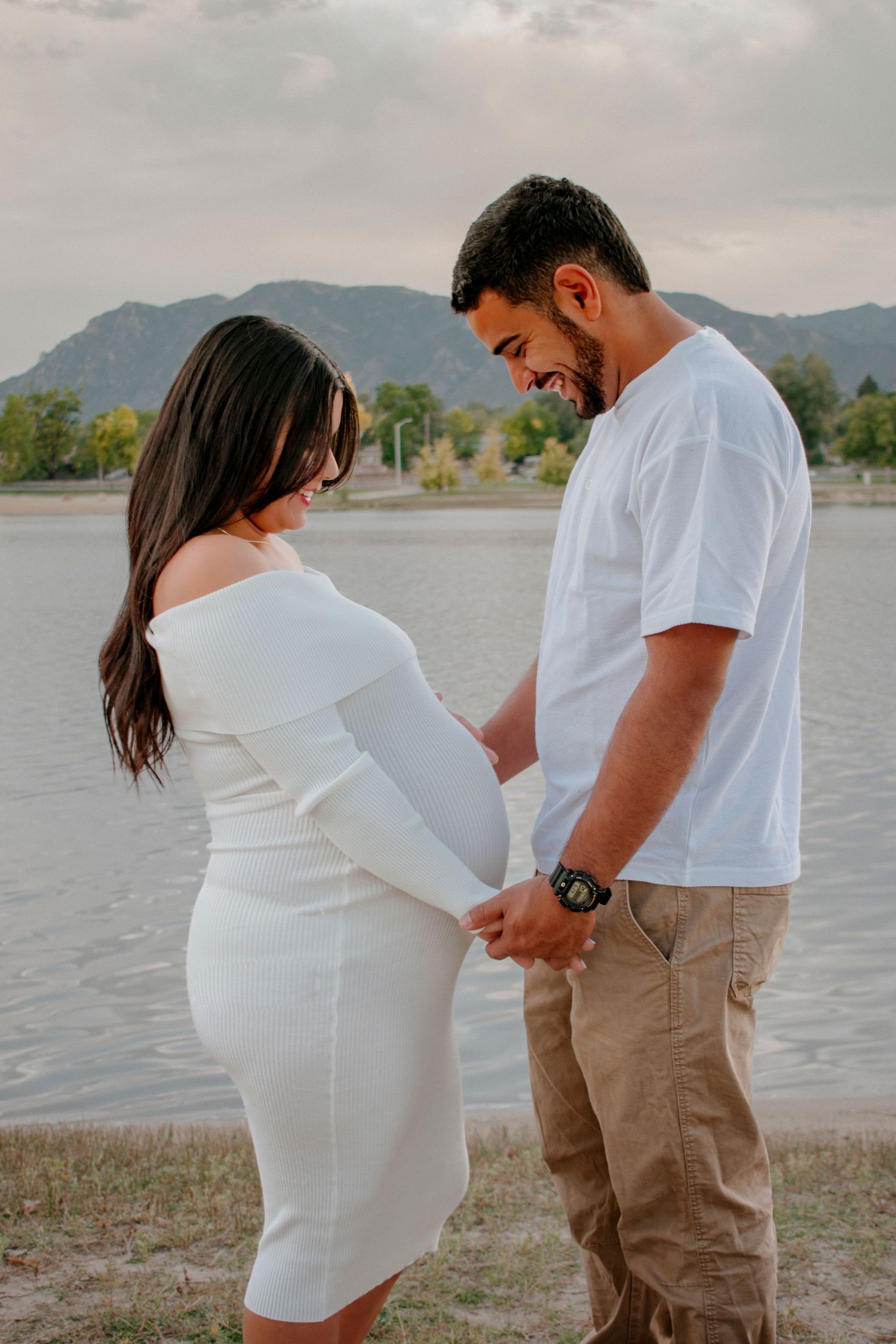 Happy couple holding hands smiling at pregnant belly near a lake with mountains in the background.