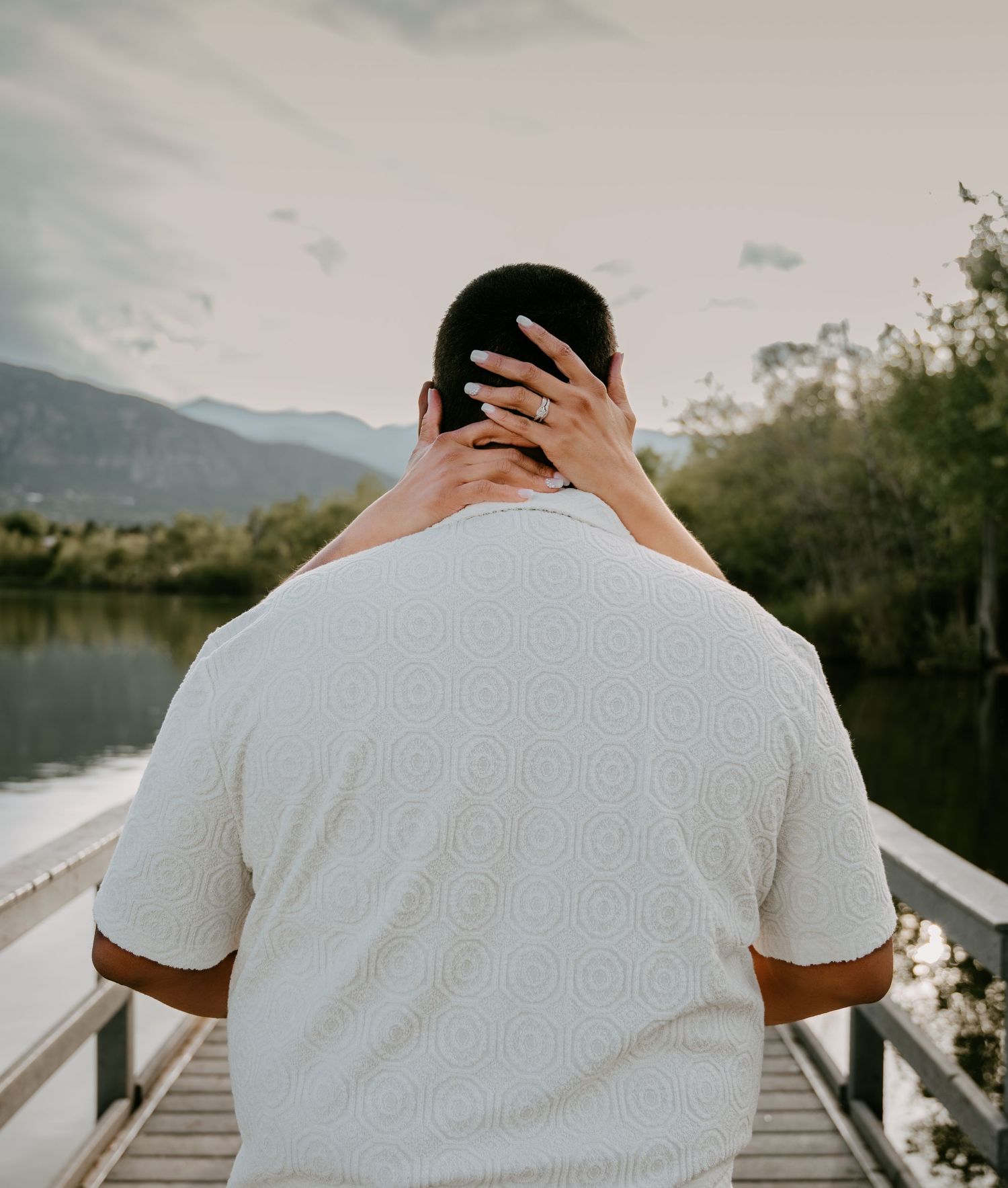 A man in a white patterned shirt stands on a dock, viewed from behind, with a woman's hand on his neck.