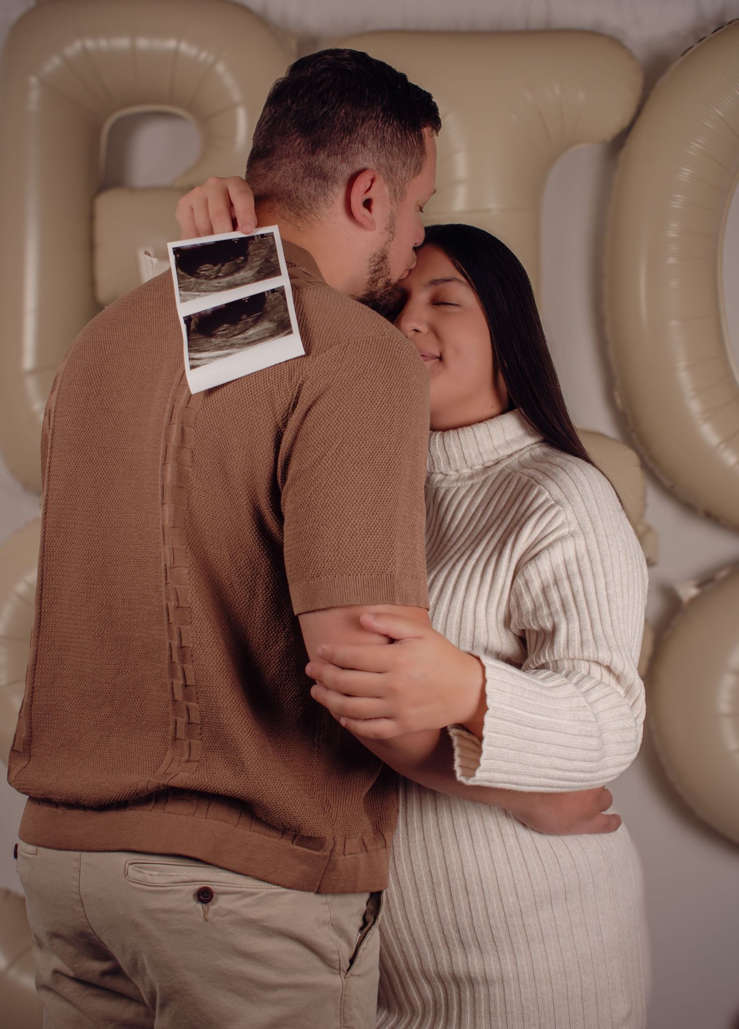 Couple embracing and holding ultrasound photos during a baby gender reveal party with balloon letters in the background.