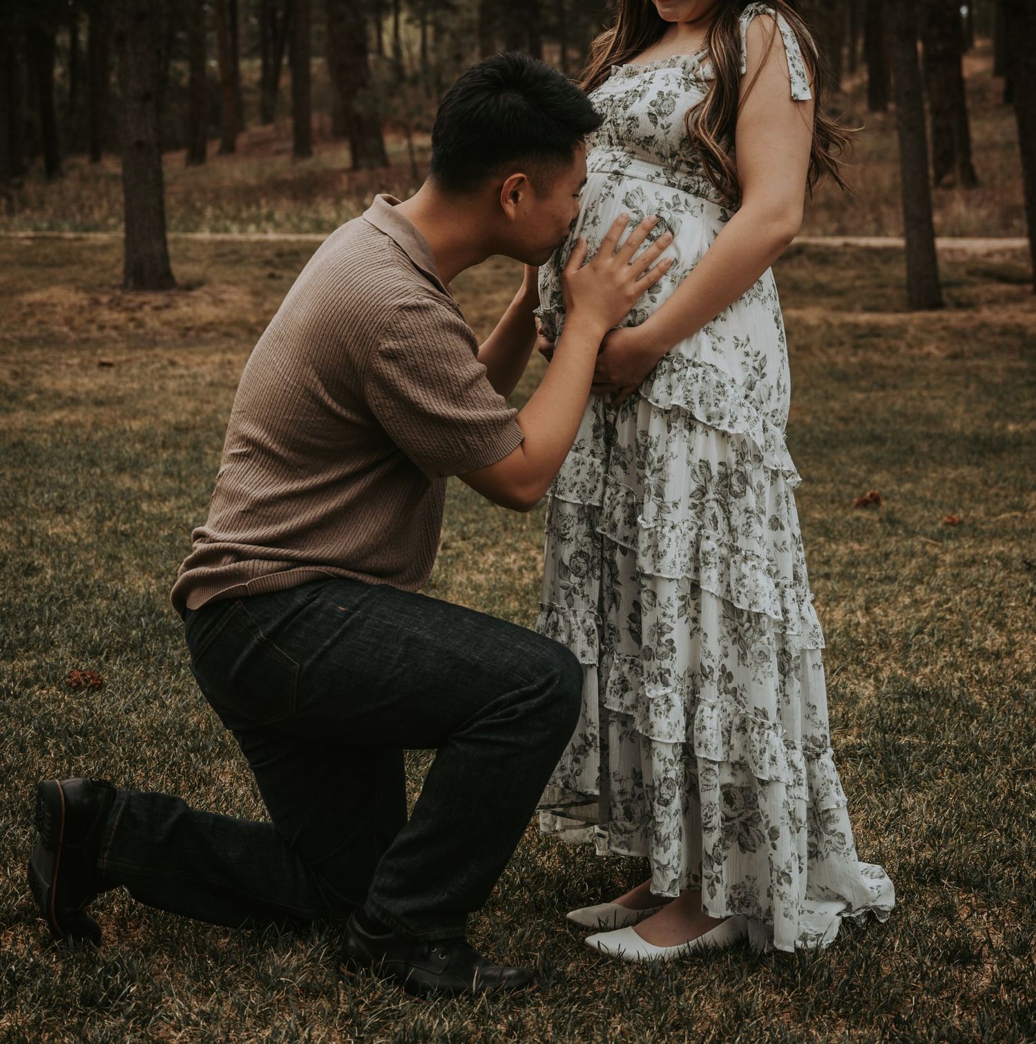 A man kneels on grass kissing a pregnant woman's belly during an outdoor maternity photo shoot in a forest.