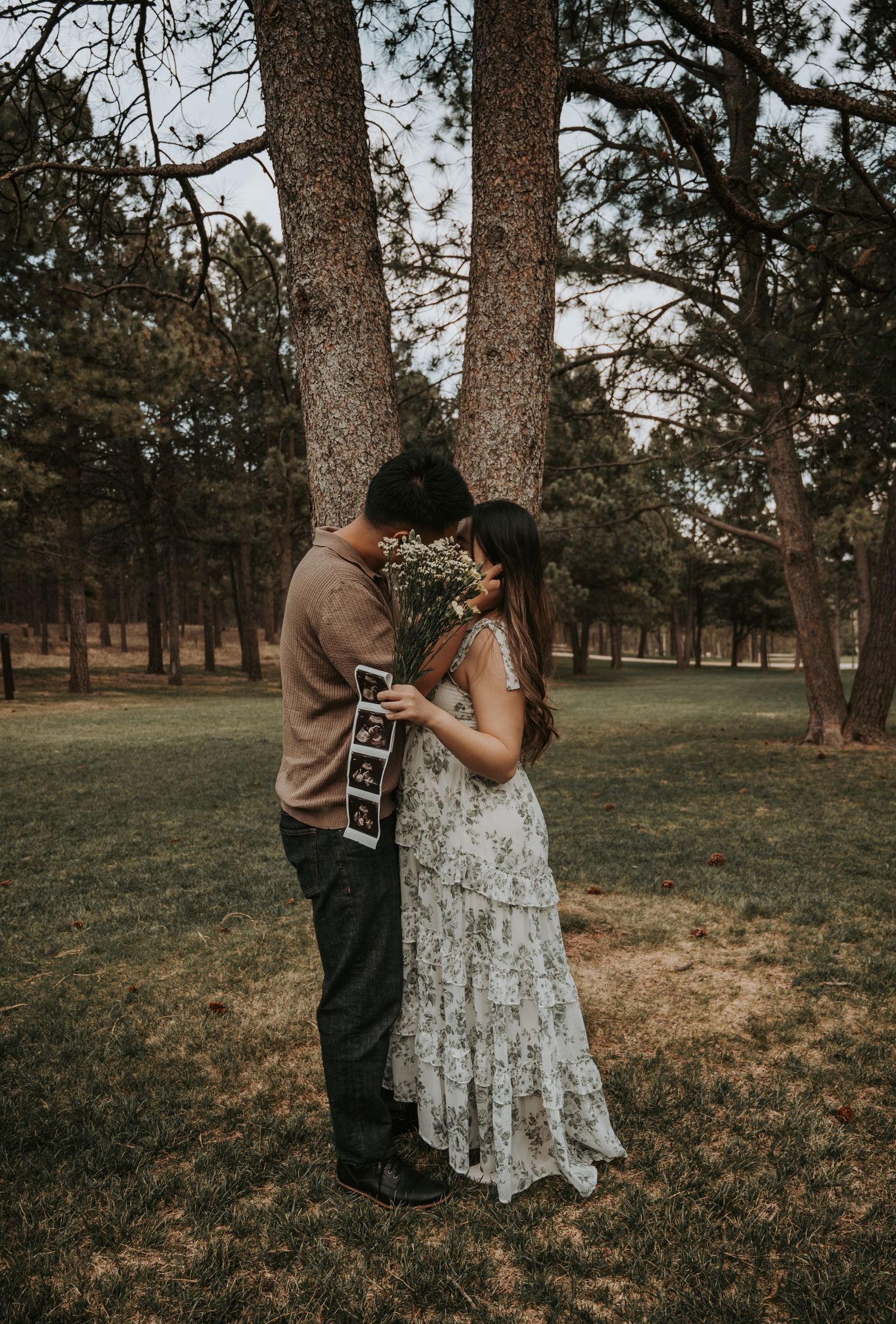 Couple sharing intimate moment in park holding wildflowers and ultrasound photos, pregnancy announcement in wooded setting.