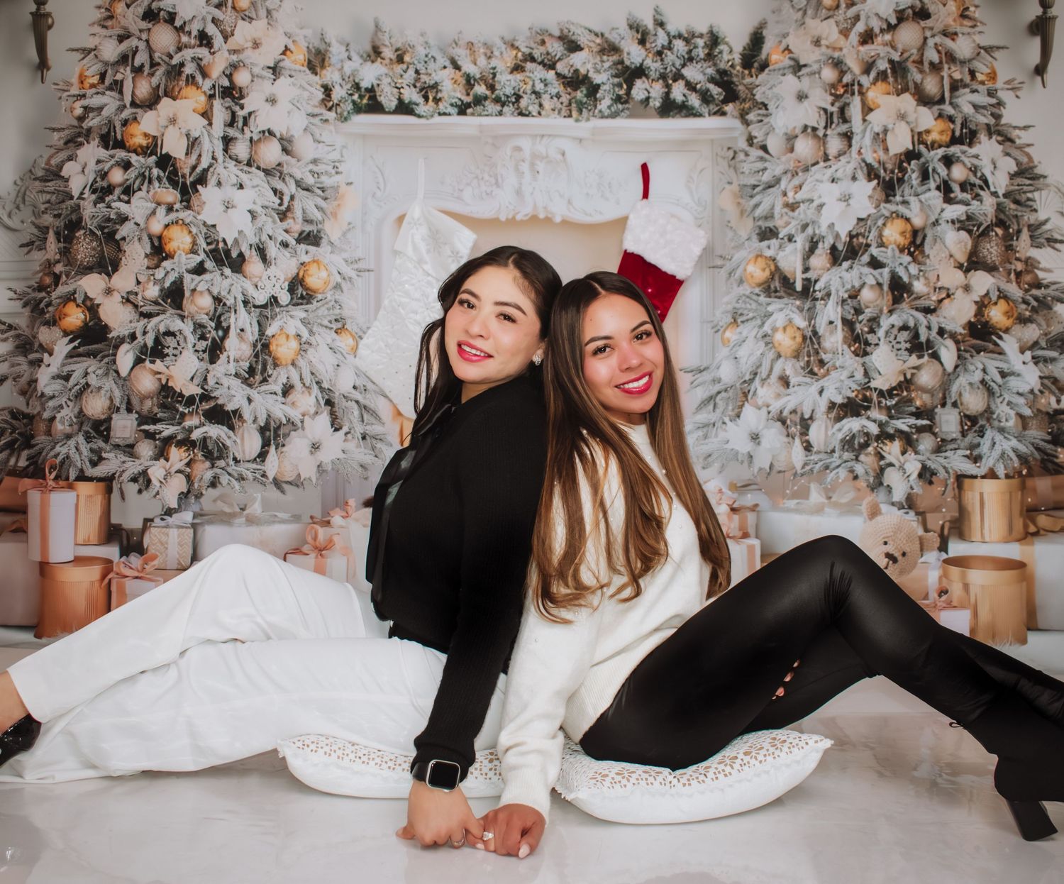 Two young women posing back-to-back in front of decorated Christmas trees with stockings and gifts.