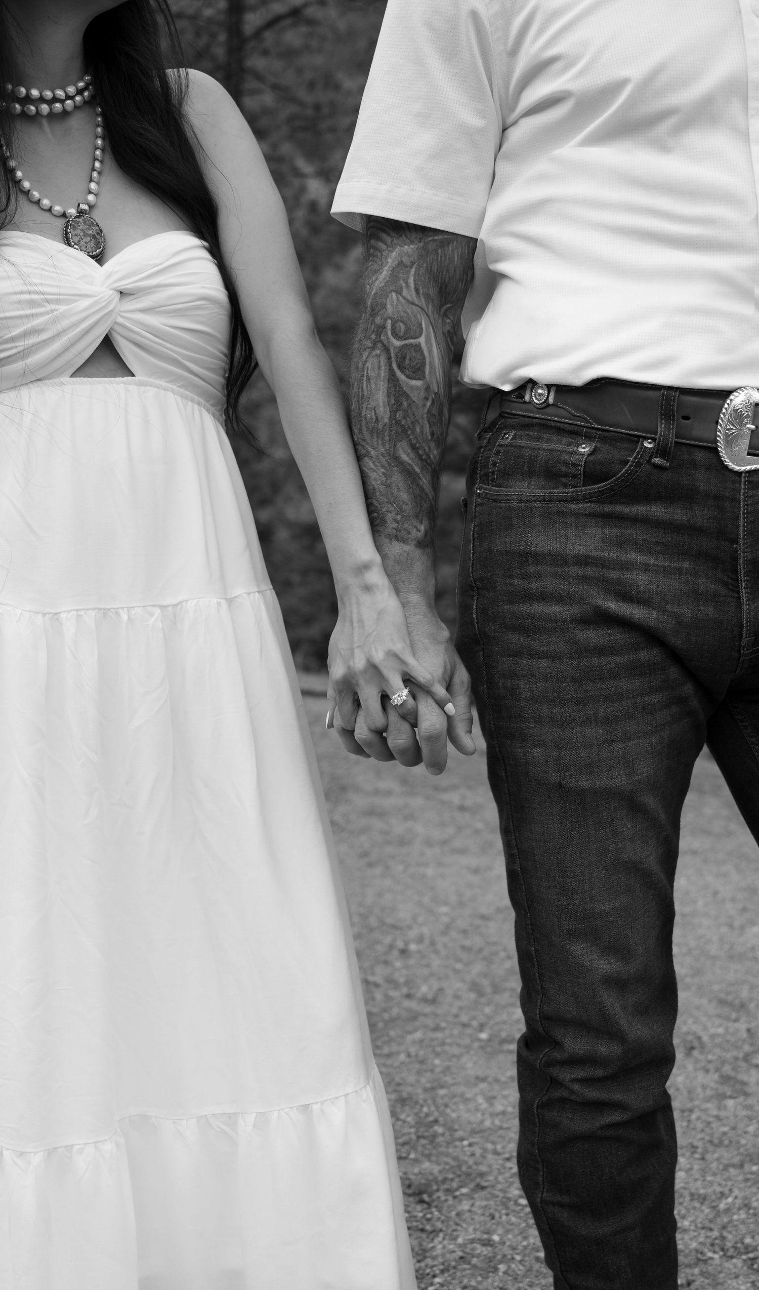 Black and white photo of a couple holding hands, woman in white dress with ring visible, man in jeans with tattoo sleeve.