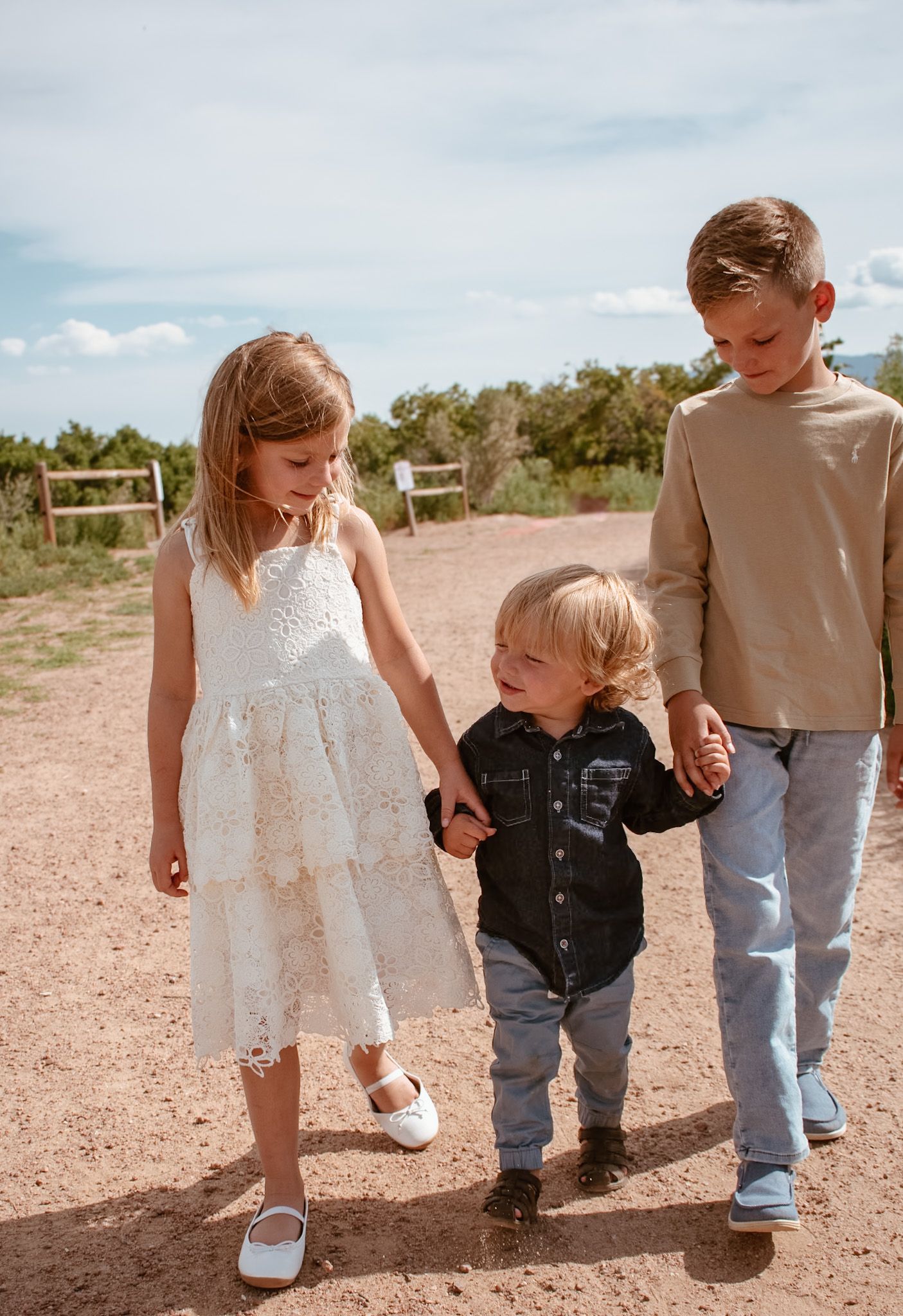 Two older siblings hold hands with a smiling toddler on a sunny dirt path outdoors.