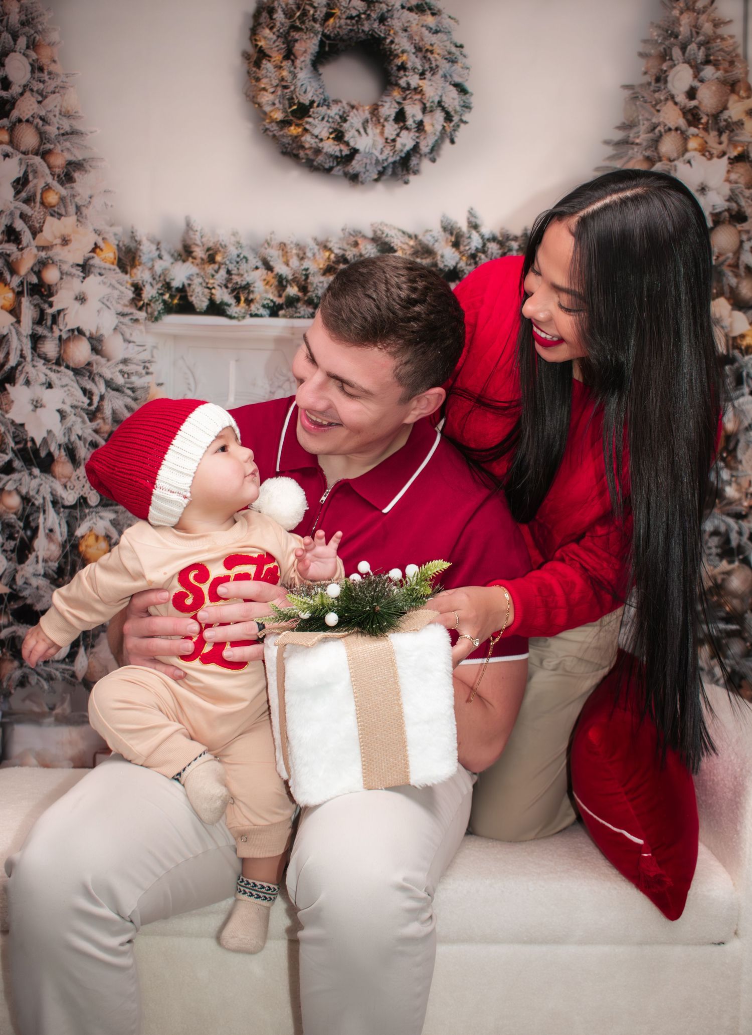Young couple in red outfits with baby in Santa hat opening Christmas gift near holiday decorations.
