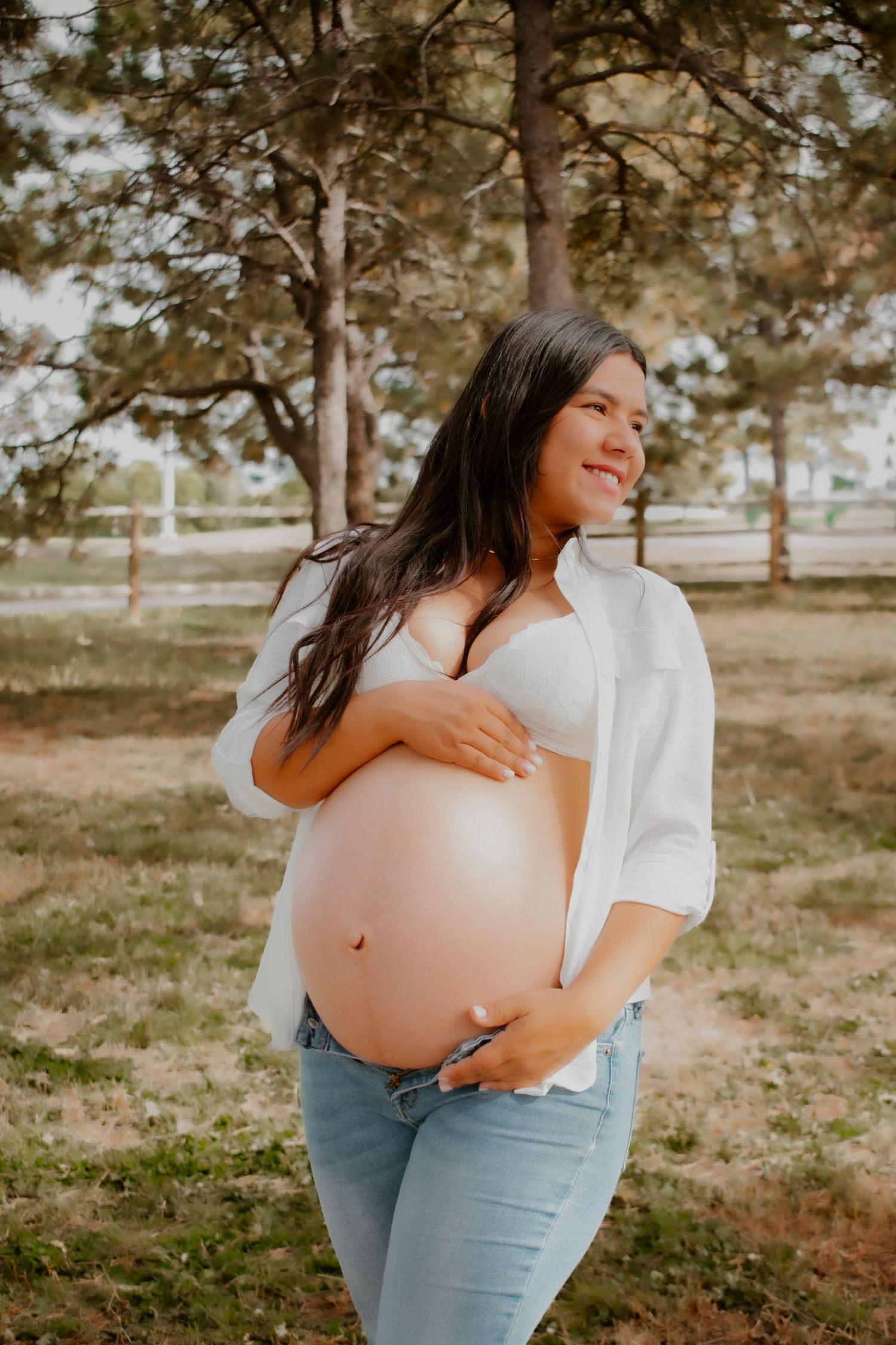 Glowing pregnant woman in white open shirt and jeans holding her bare belly while smiling in a park.