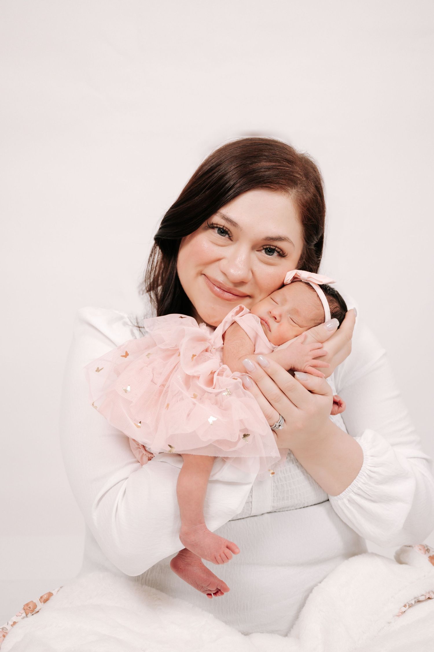 A smiling brunette woman in white holds a sleeping newborn baby dressed in a pink outfit against a light background.