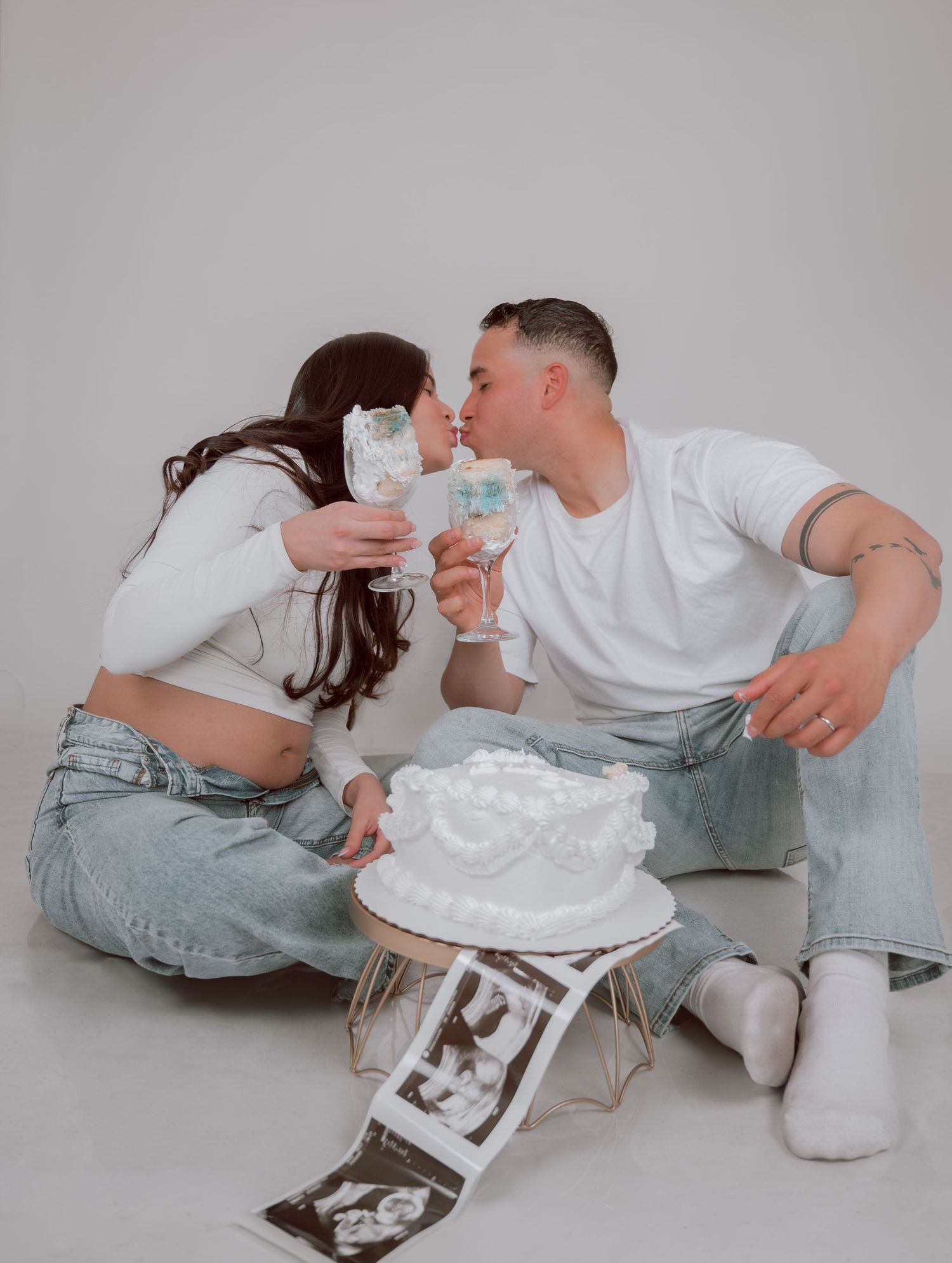 Pregnant couple kissing at maternity photoshoot with white cake and ultrasound photos on white background.