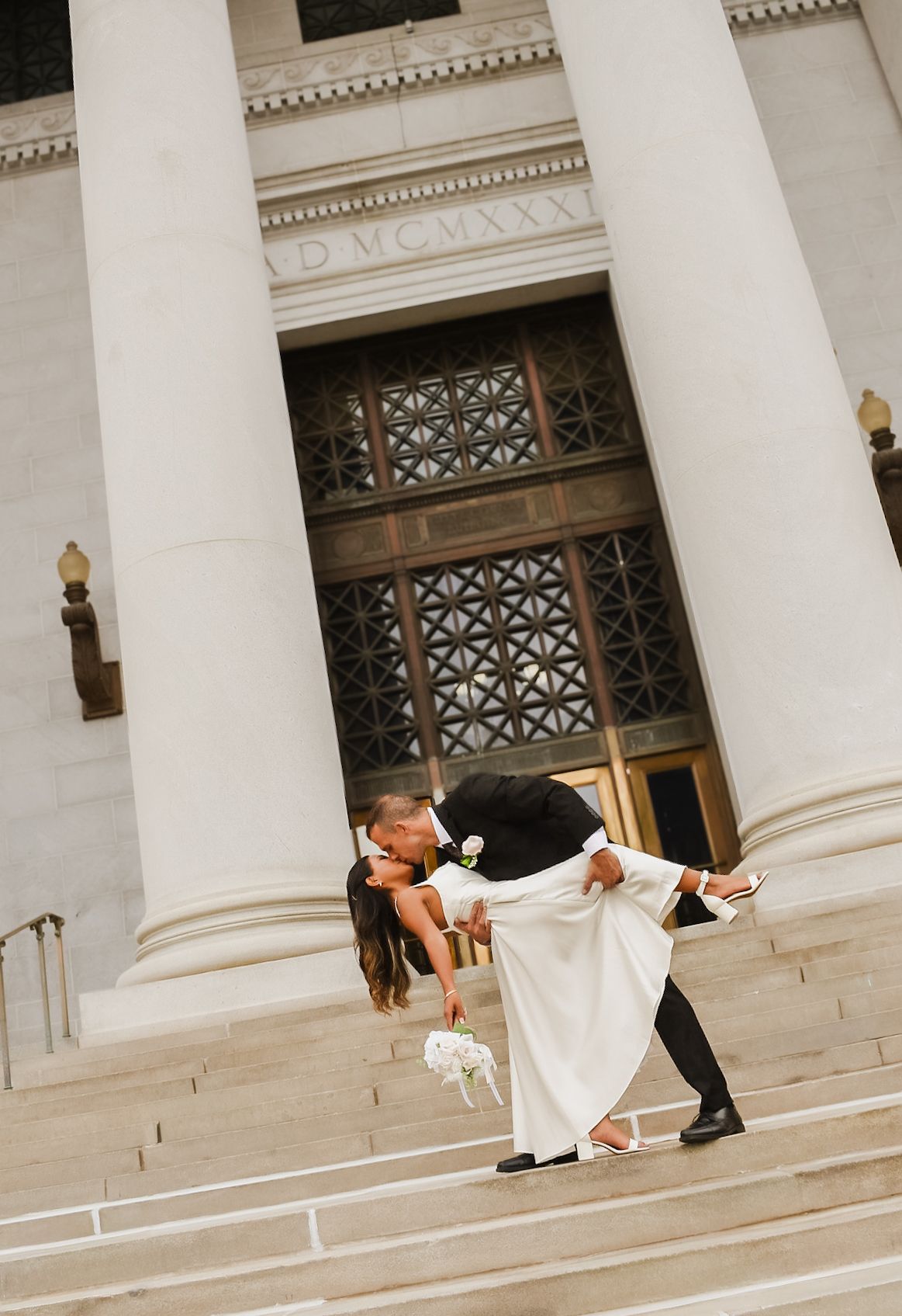 Groom dipping bride in wedding kiss on steps of grand building with tall white columns.