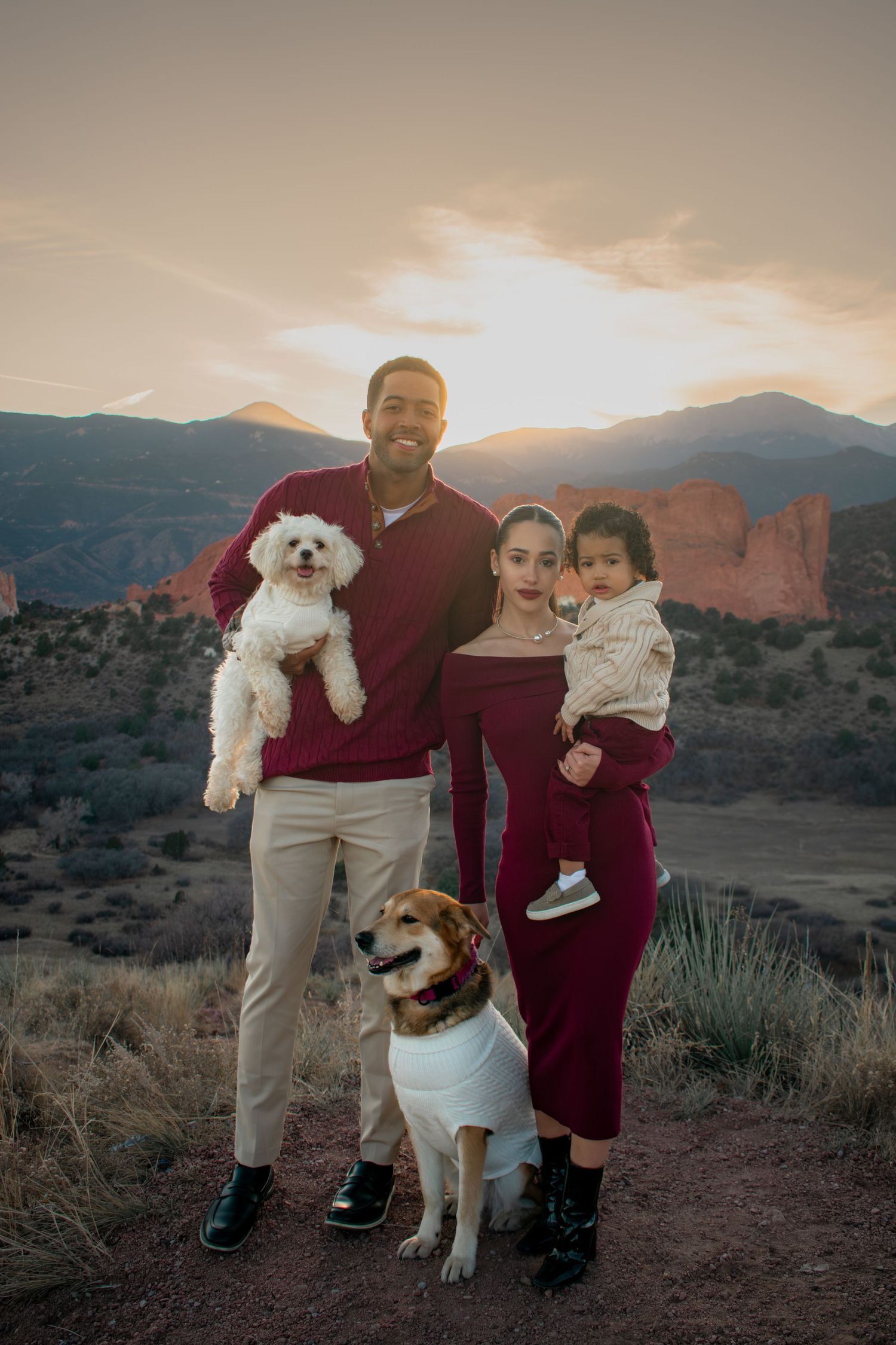 Family portrait at Garden of the Gods with two adults, toddler, and two dogs at sunset with mountain backdrop.