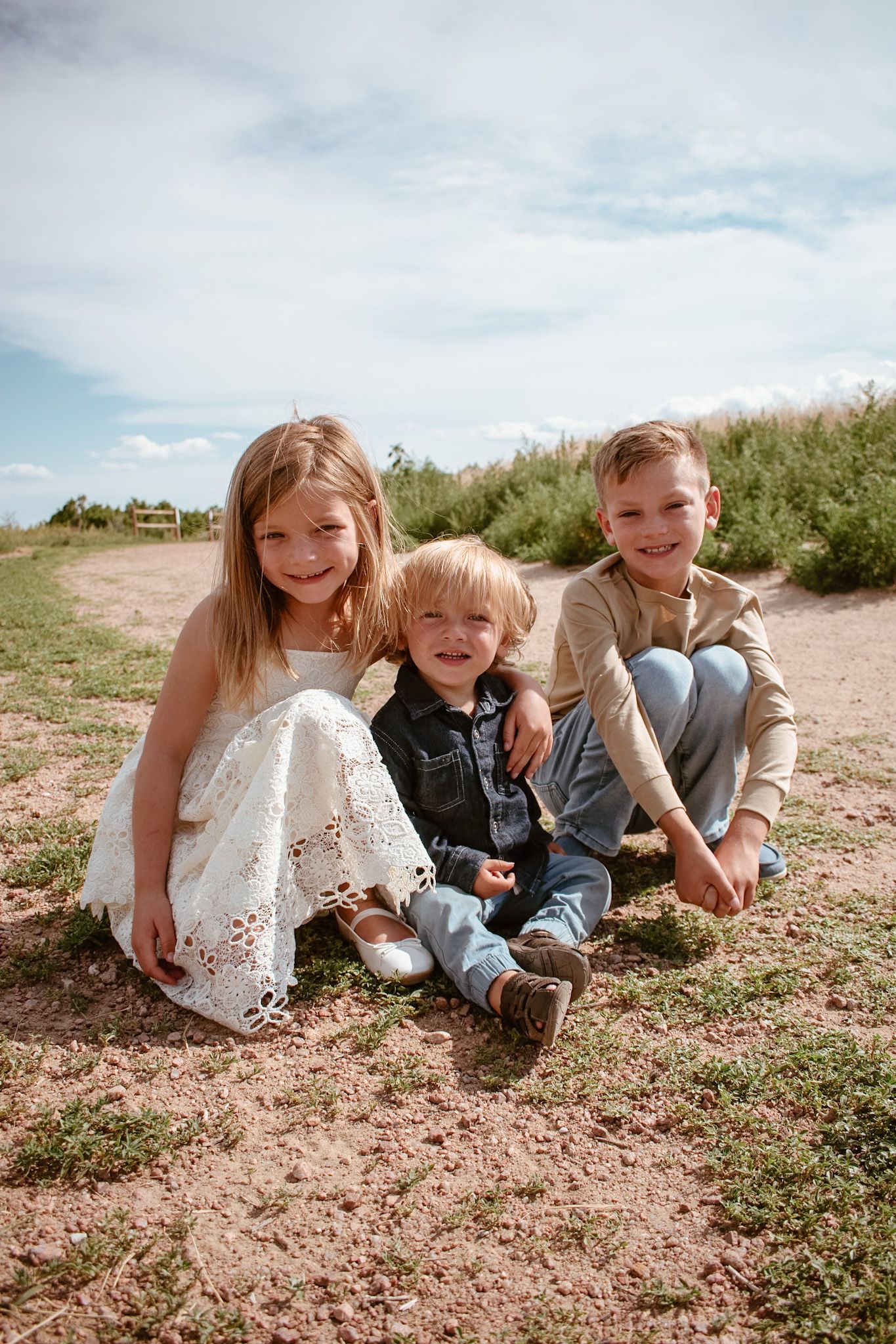 Three young siblings sit together smiling on a dirt path under a partly cloudy sky.