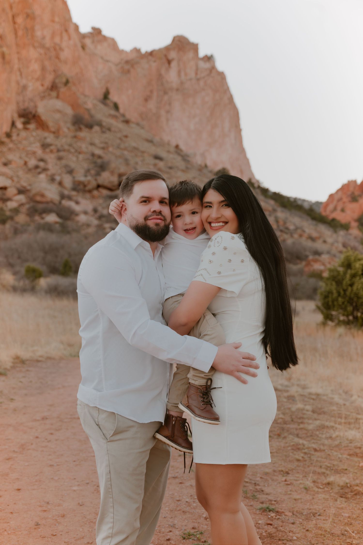 A happy family of three poses outdoors at Garden of the Gods, wearing white outfits against red rock formations.
