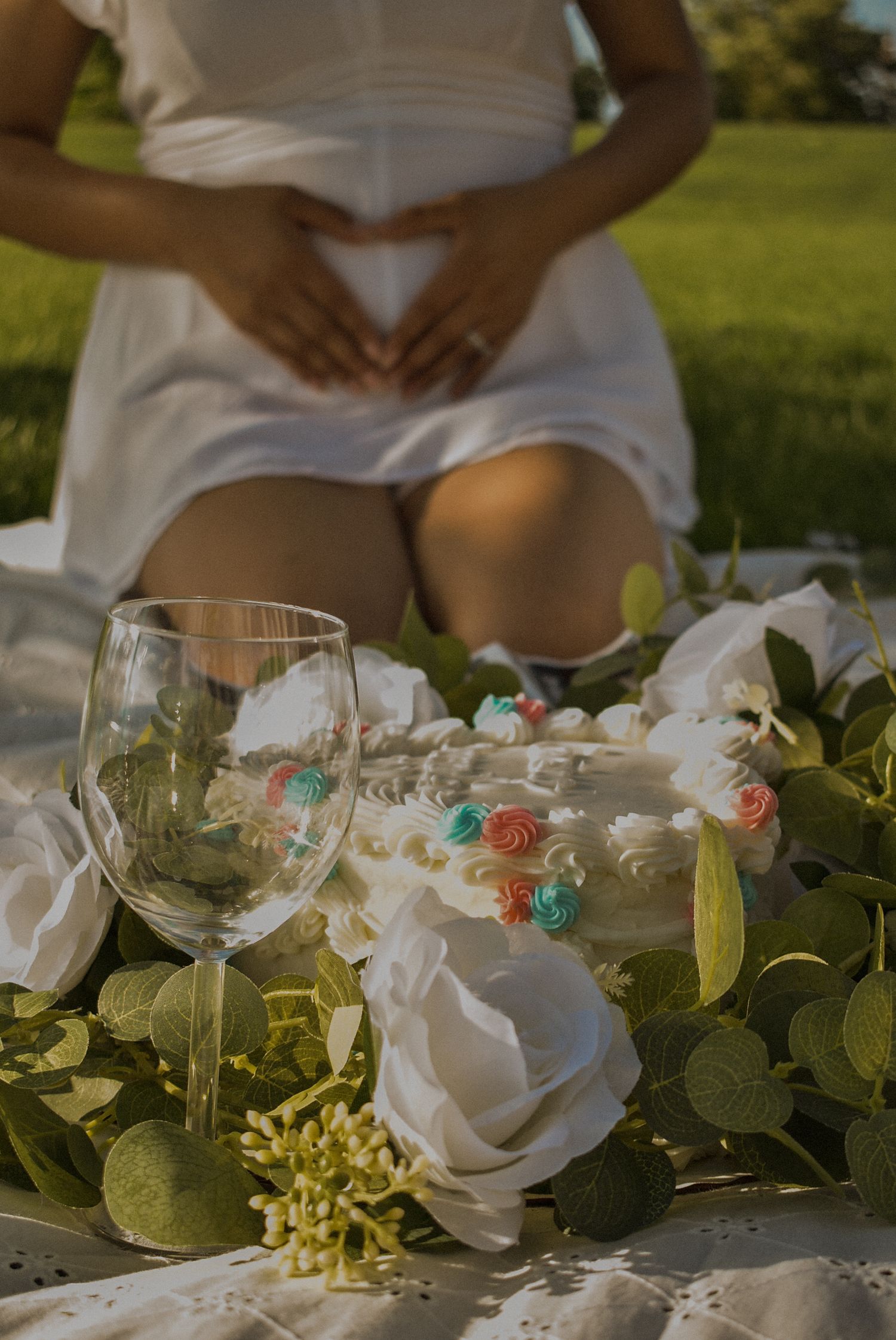 Pregnant woman in white dress kneeling outdoors with wine glass and white floral arrangement in foreground.