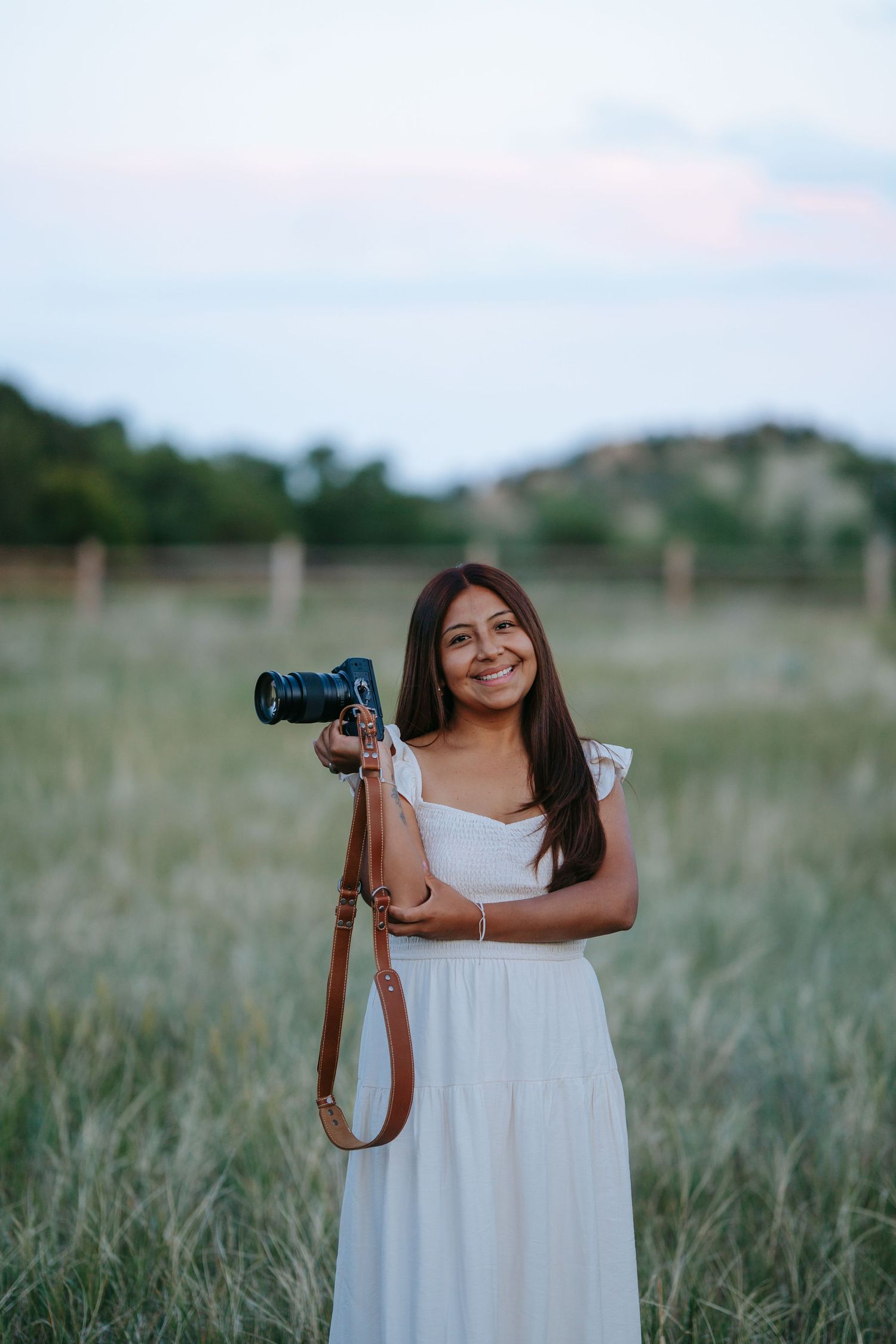 Young woman in white dress holding DSLR camera with brown strap, smiling in a scenic grassy field at dusk.