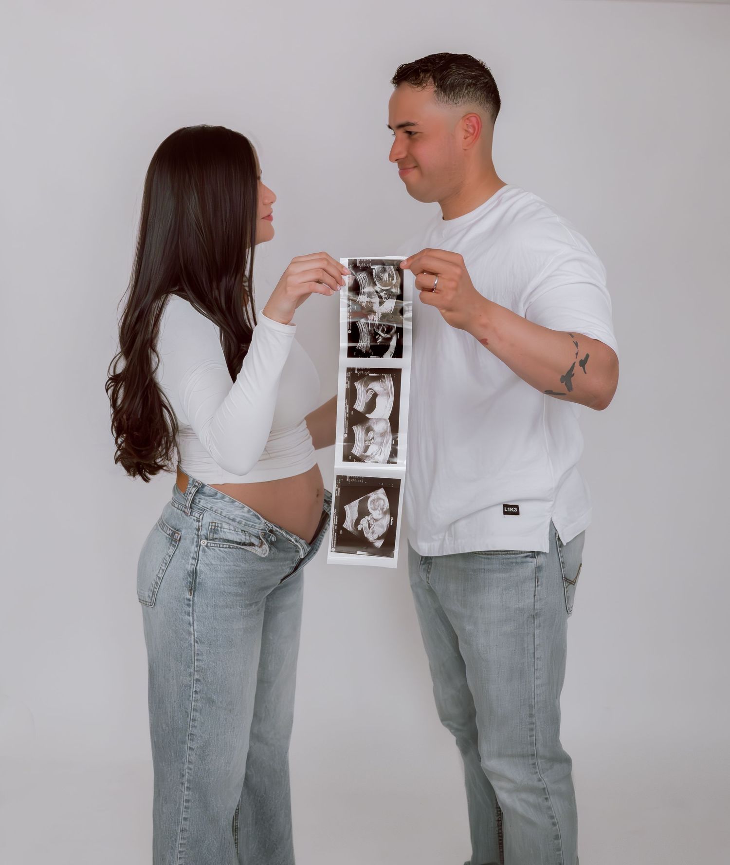 Couple holding ultrasound photos together, pregnancy announcement, wearing white tops and jeans against white background.