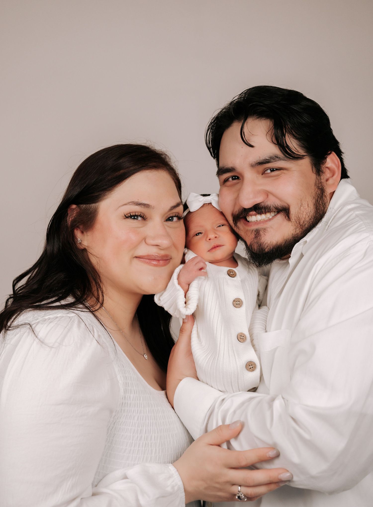 A smiling couple in white outfits holds their newborn baby dressed in white against a light background.