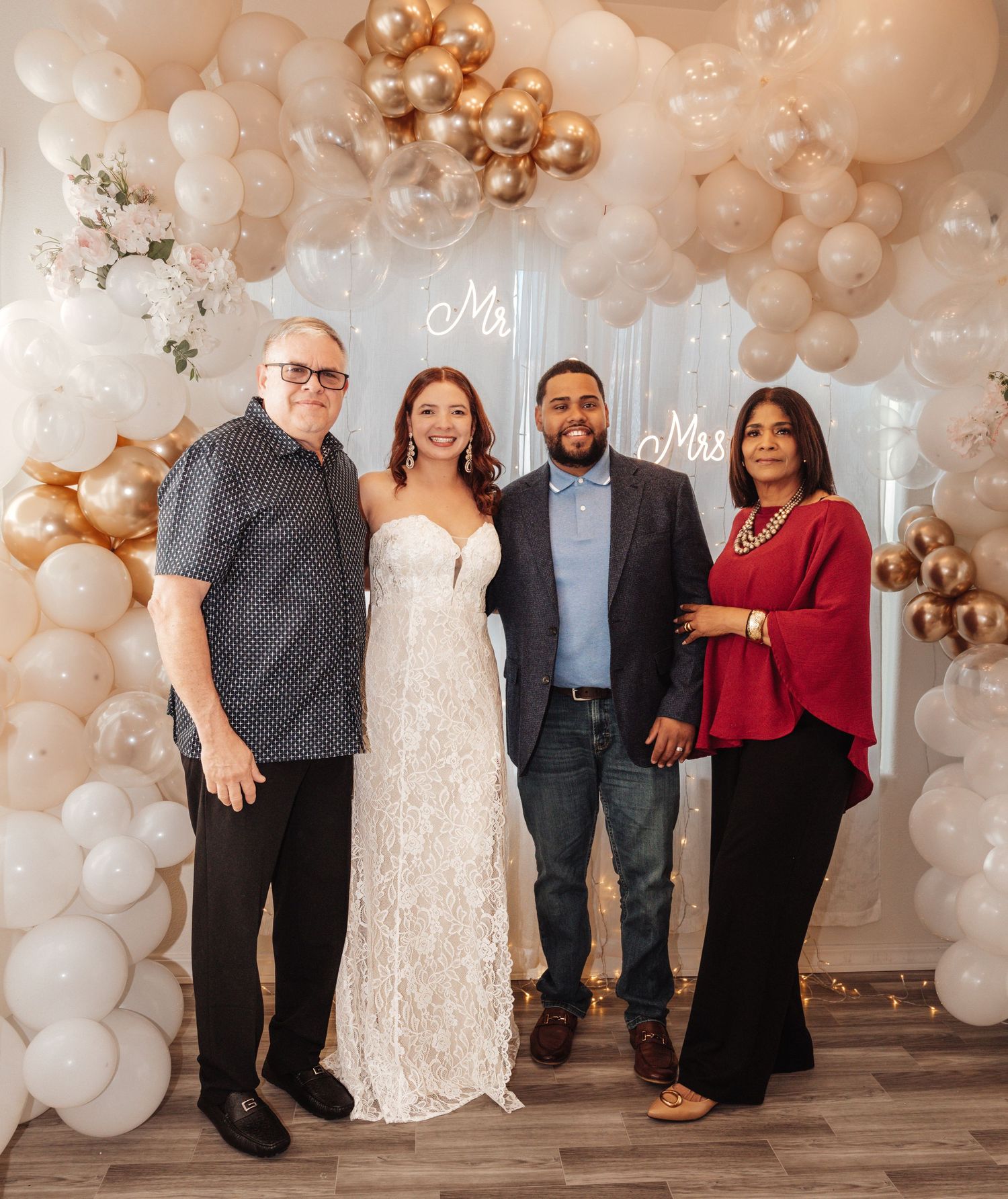 Four people pose together at a wedding celebration with white and gold balloon arch backdrop and Mr. & Mrs. neon sign.