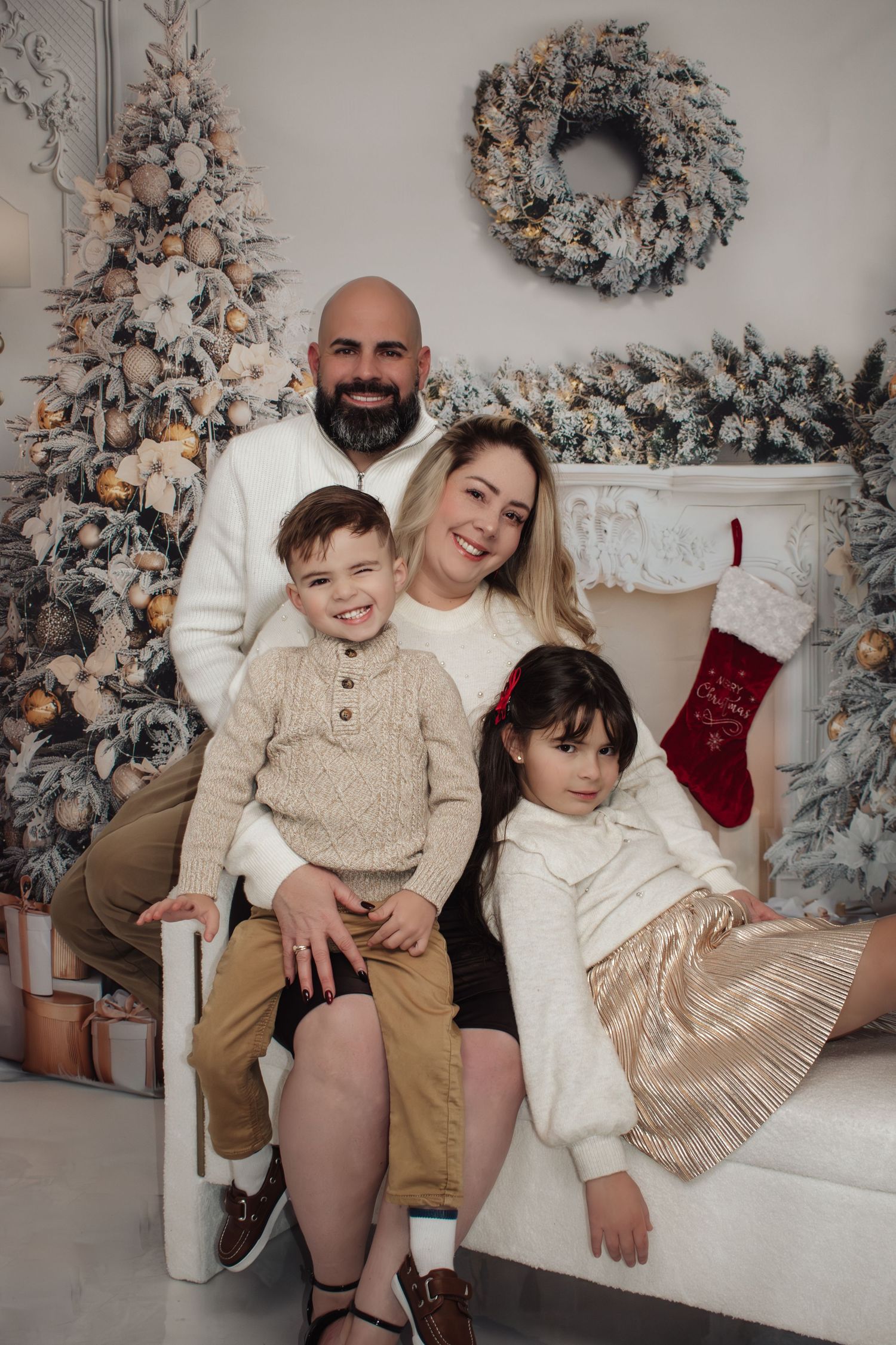 Bald bearded father, blonde mother, and two children smiling in front of Christmas tree and fireplace.
