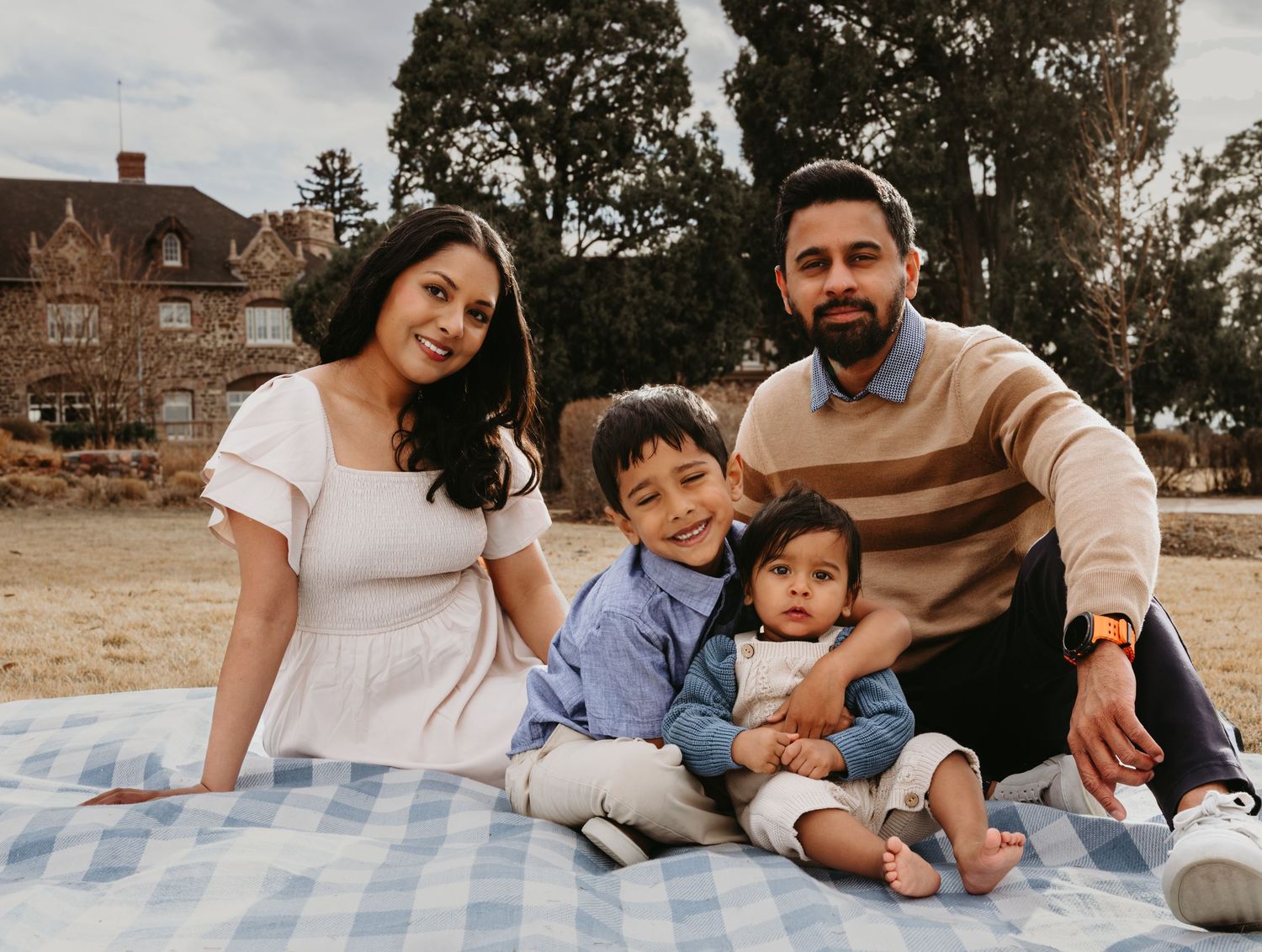 Happy South Asian family of four smiling together on a picnic blanket outdoors near a historic building.