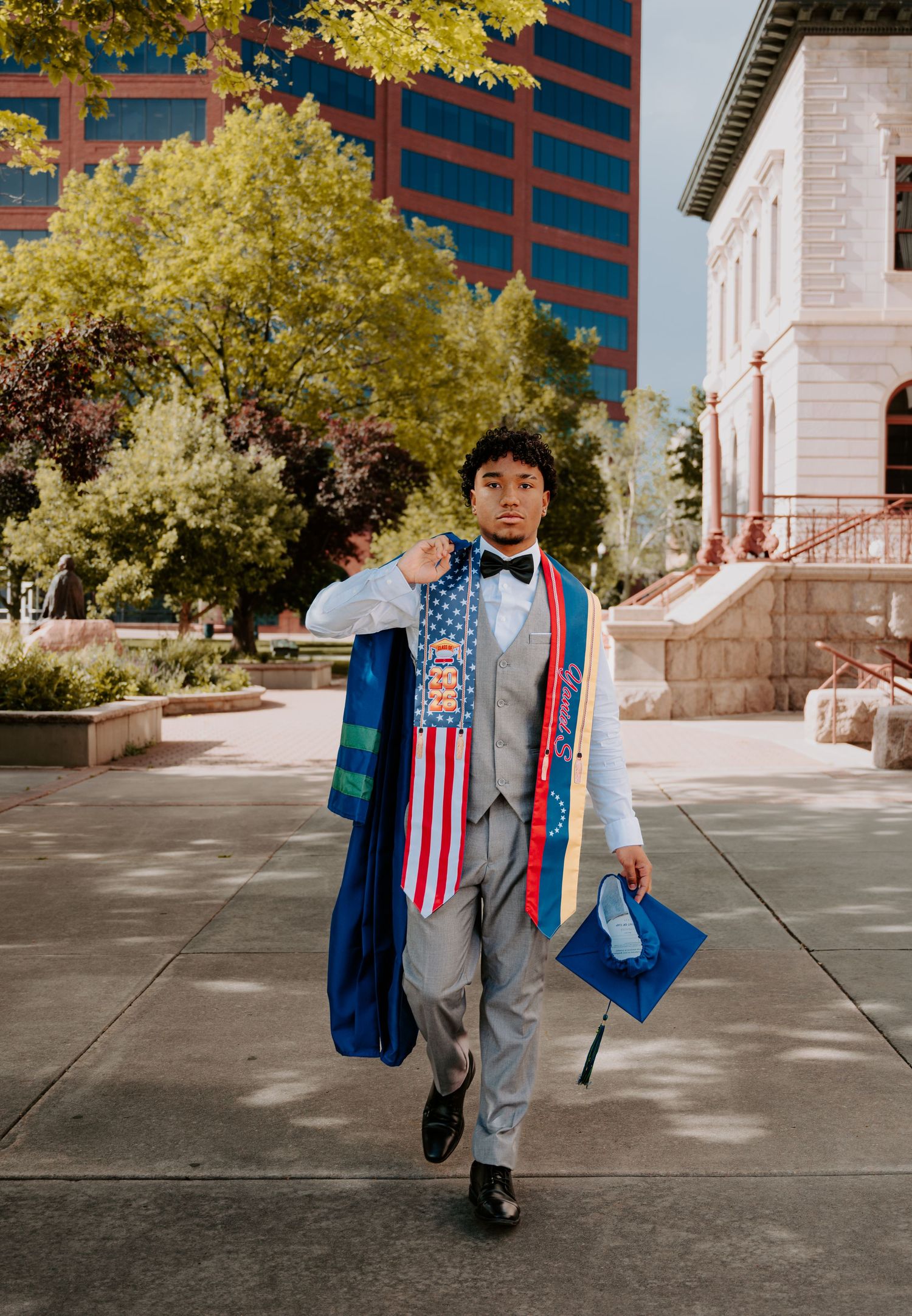 Young man in gray suit with graduation stoles and cap walks confidently outdoors after commencement ceremony.