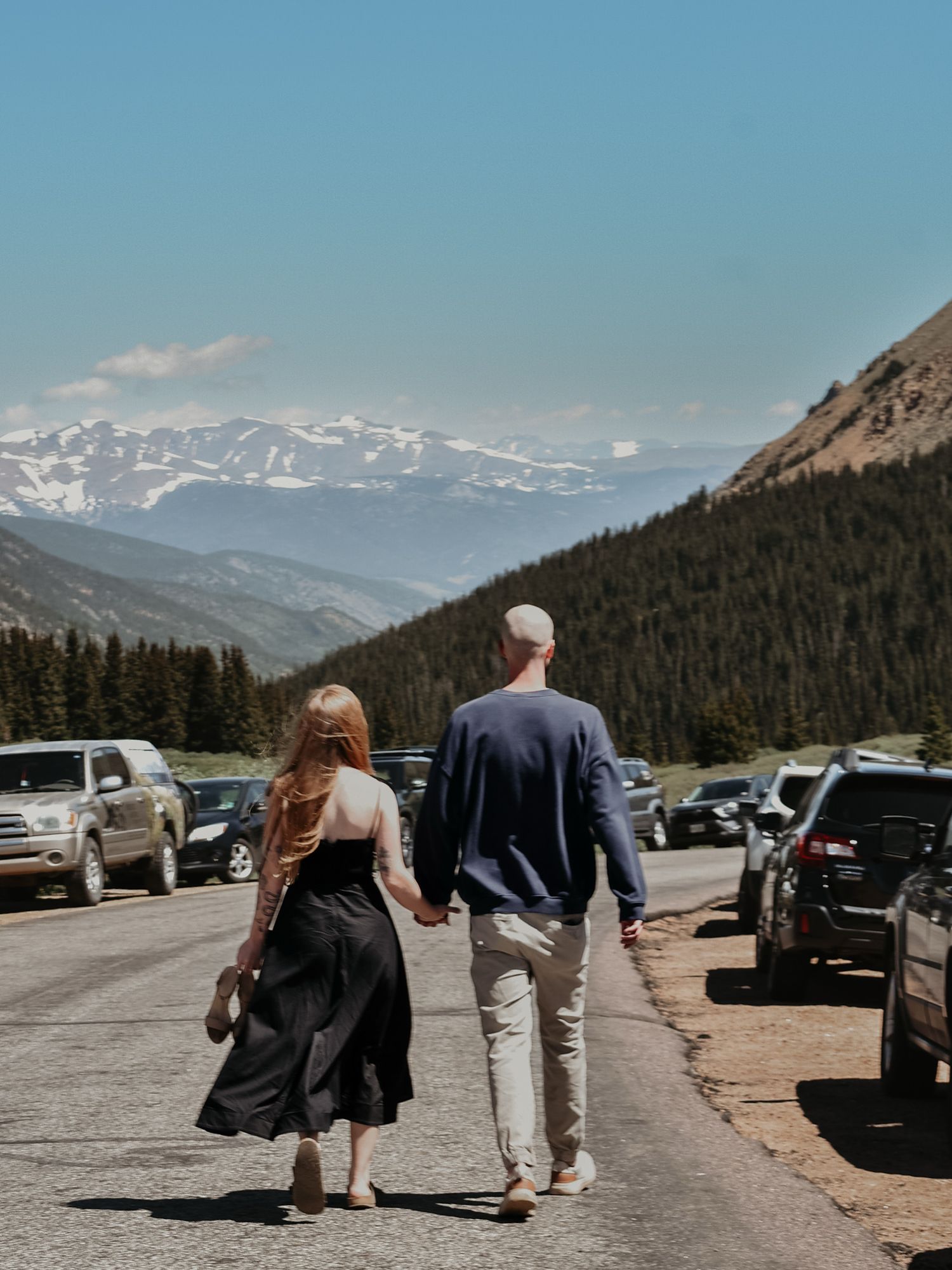 Couple walking hand in hand through a mountain parking lot with snow-capped peaks in the background.