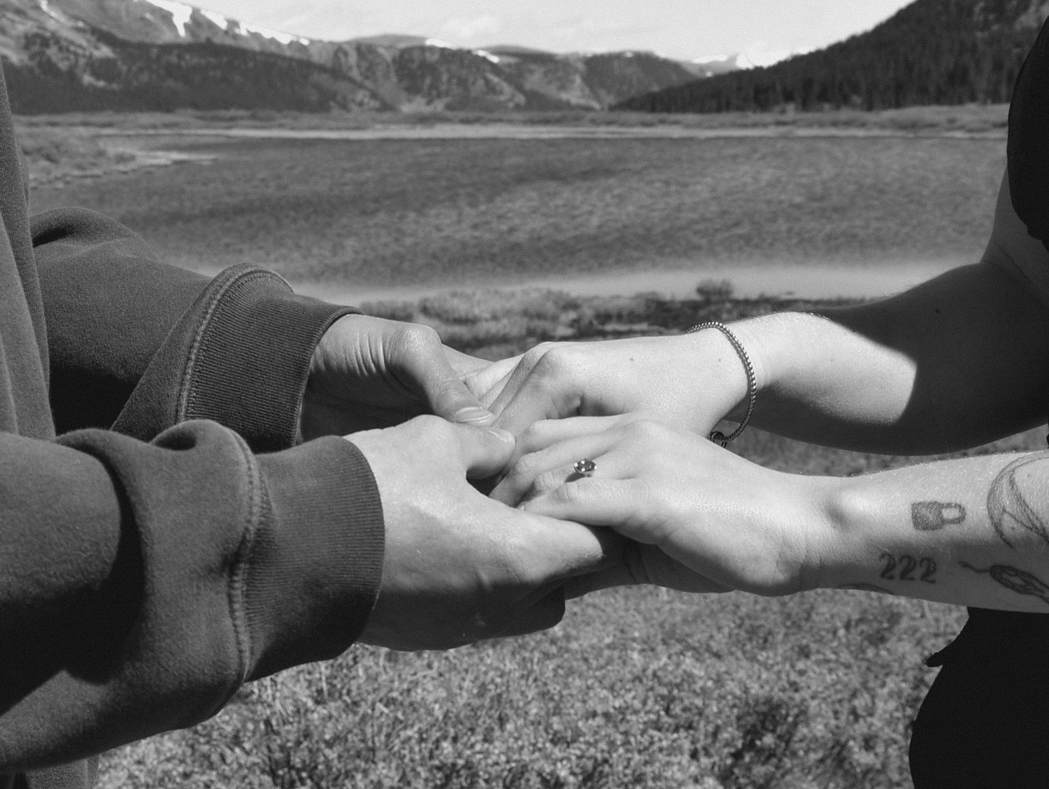 Black and white close-up of couple holding hands showing engagement ring with mountain landscape behind them.
