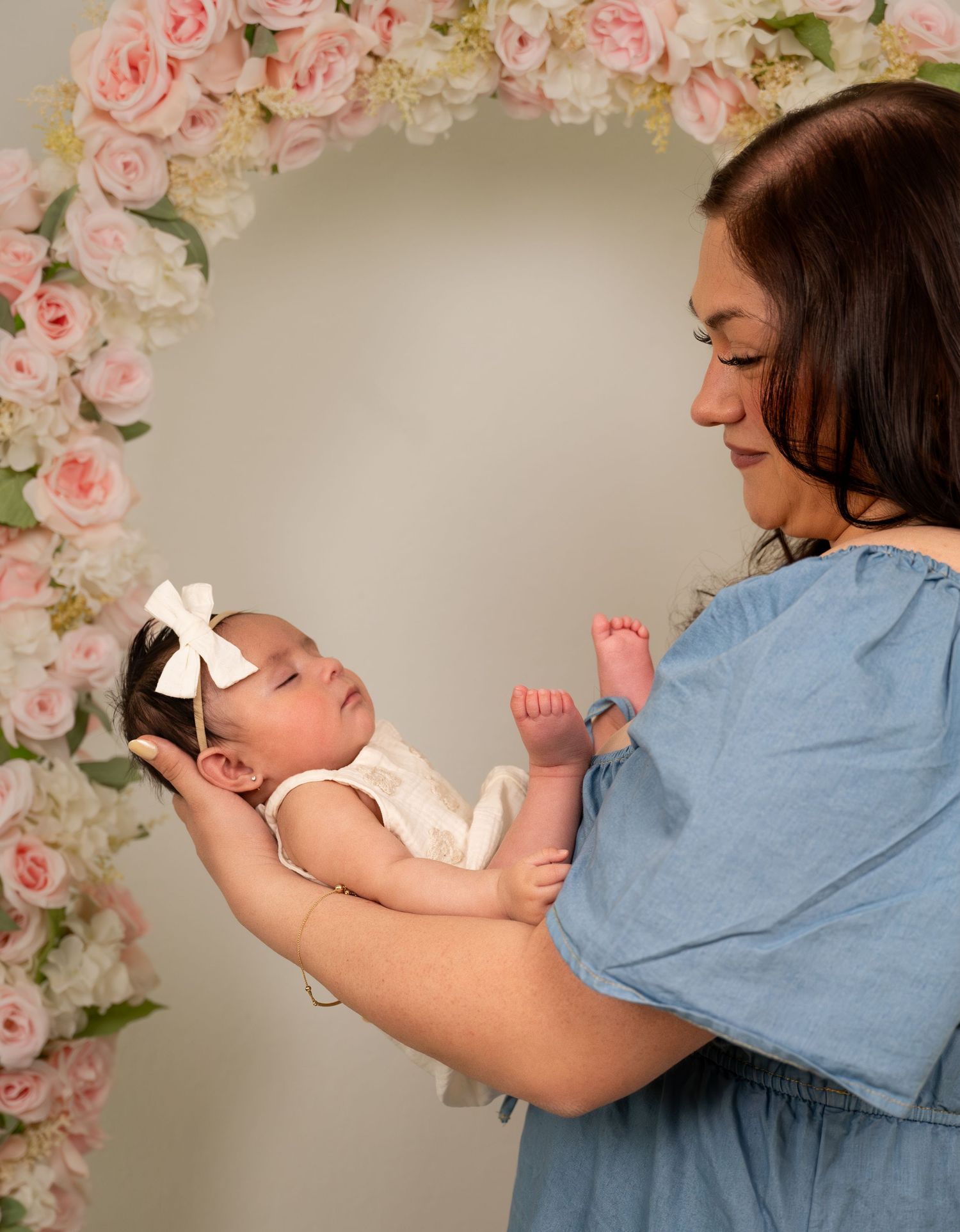 Mother in blue dress holds newborn baby with white bow headband in front of pink floral arch backdrop.