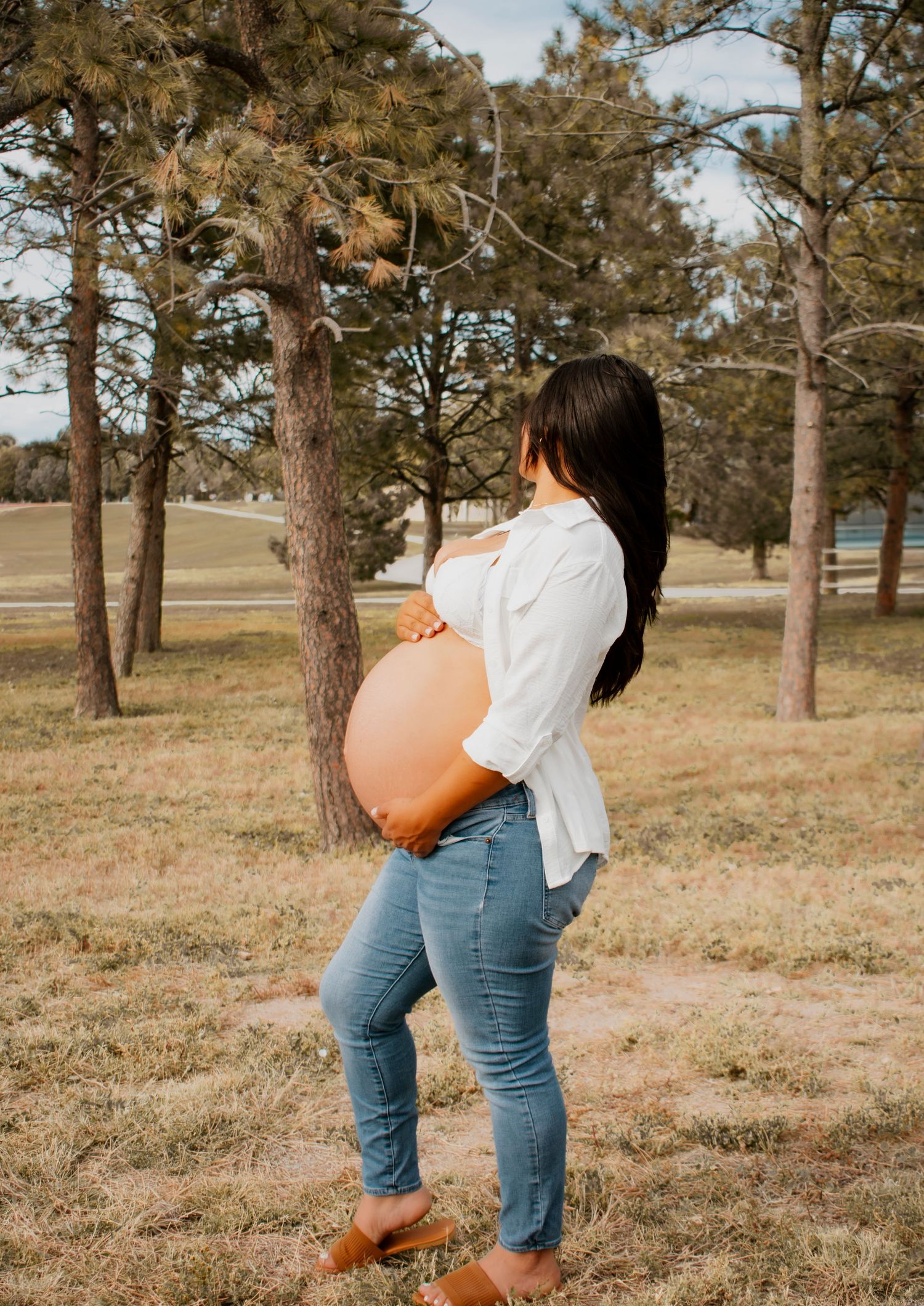 Pregnant woman with dark hair in white shirt and jeans showing profile of her baby bump among trees.