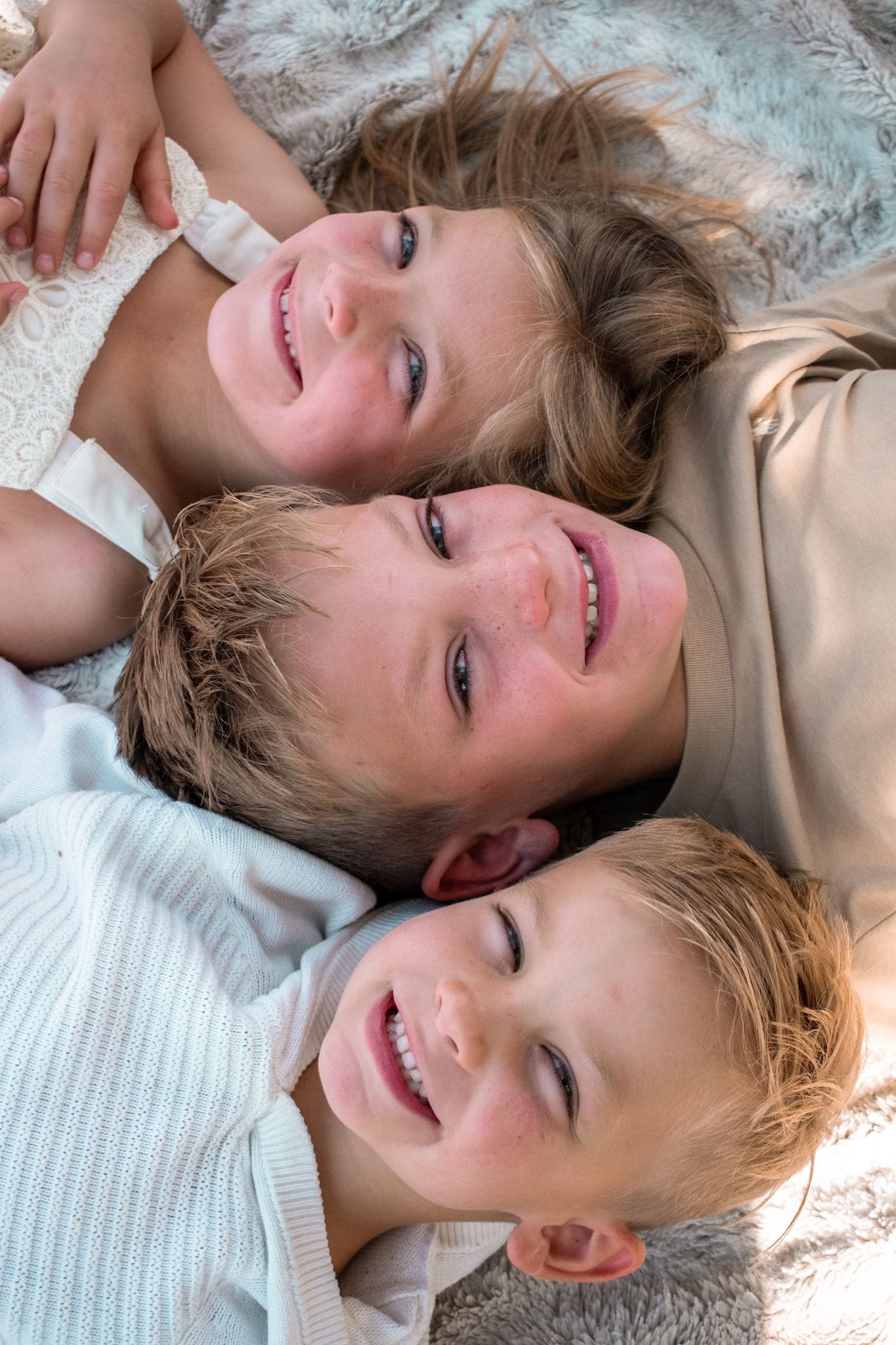 Three smiling children lying together on a soft blanket, looking up at the camera in a heartwarming sibling portrait.
