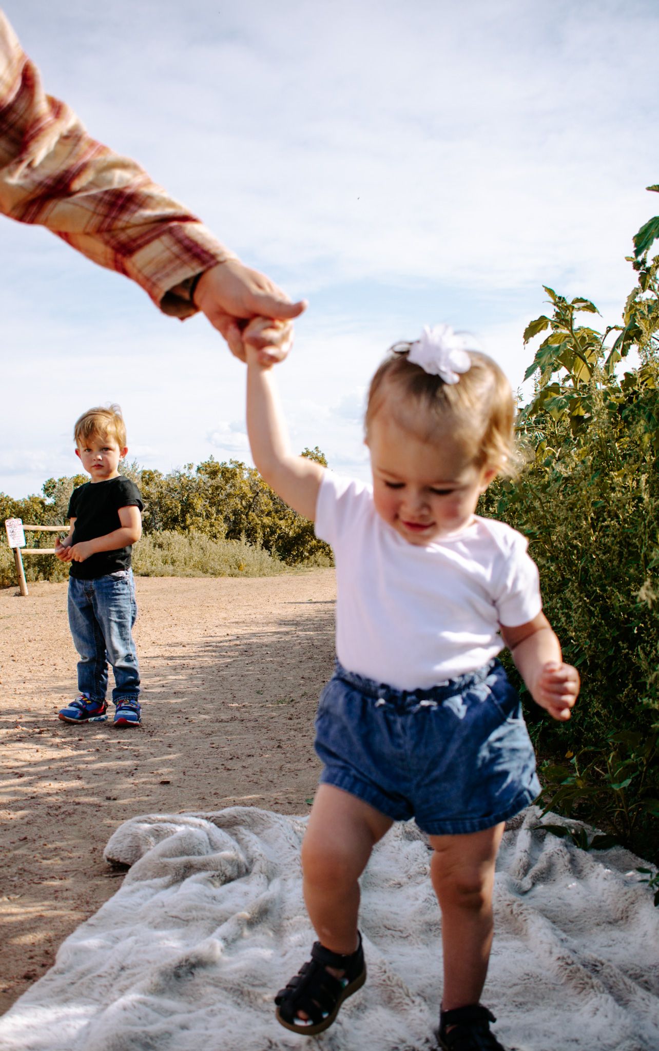A toddler girl with a white bow holds an adult's hand on a sunny outdoor path.
