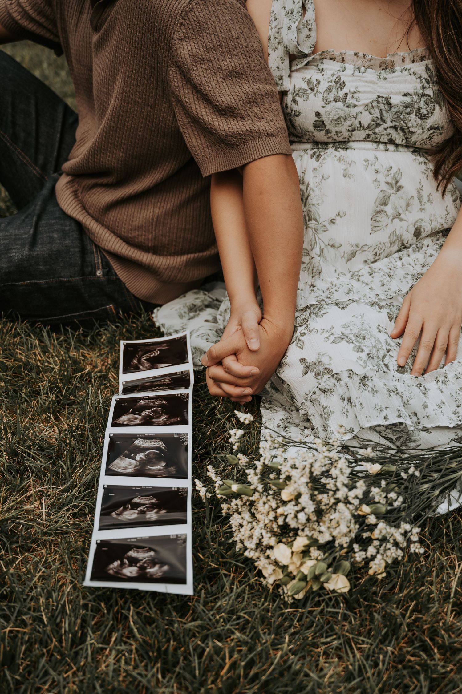 Couple holding hands on grass with ultrasound photos and white wildflowers in a pregnancy announcement photo.