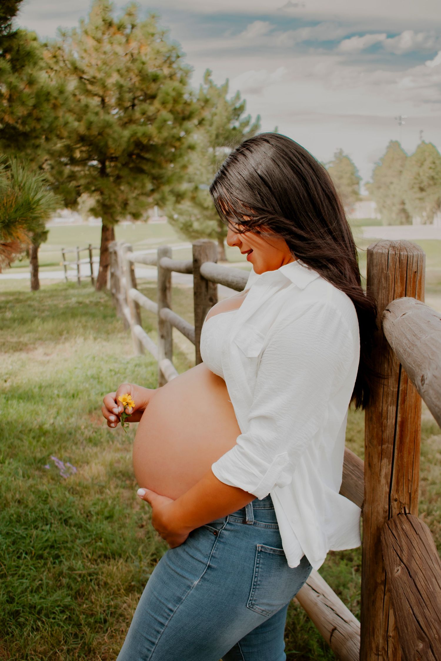 Pregnant woman in white shirt and jeans leaning on wooden fence holding a small flower over her bump.