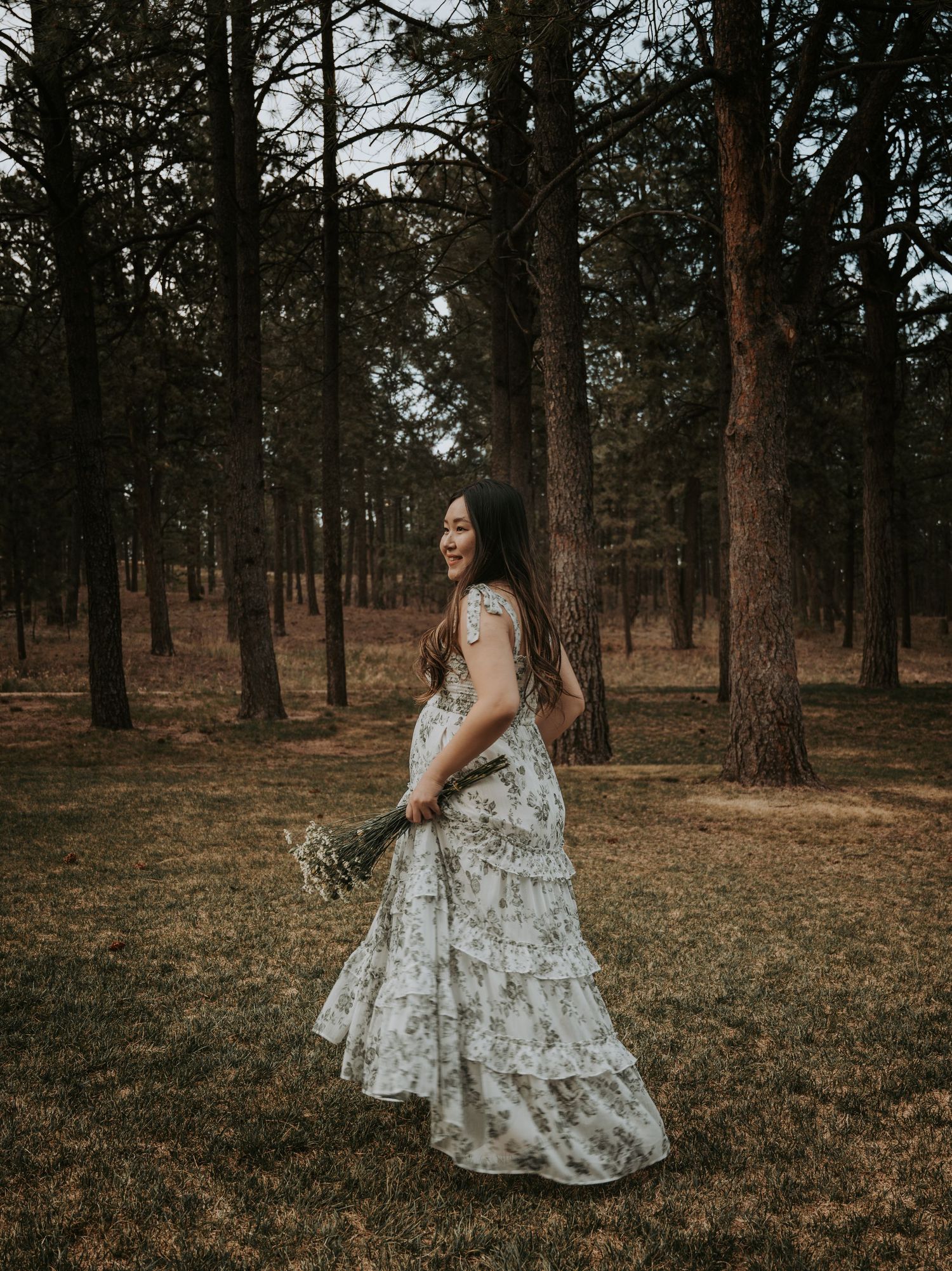 A woman in a floral maxi dress twirls while holding wildflowers in a serene pine forest setting.