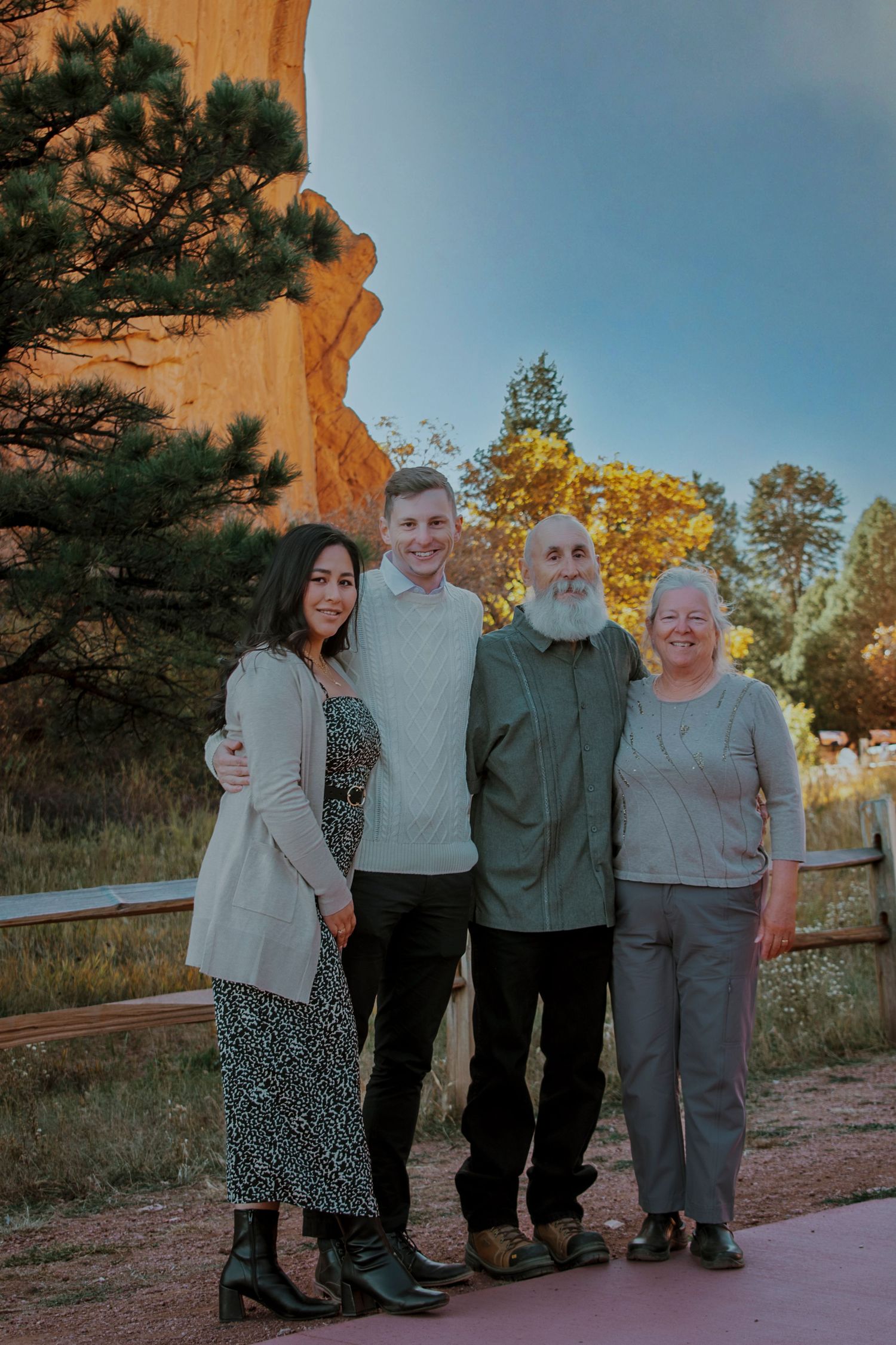 Family of four smiling together outdoors at Garden of the Gods, Colorado, with red rock formations and trees in background.