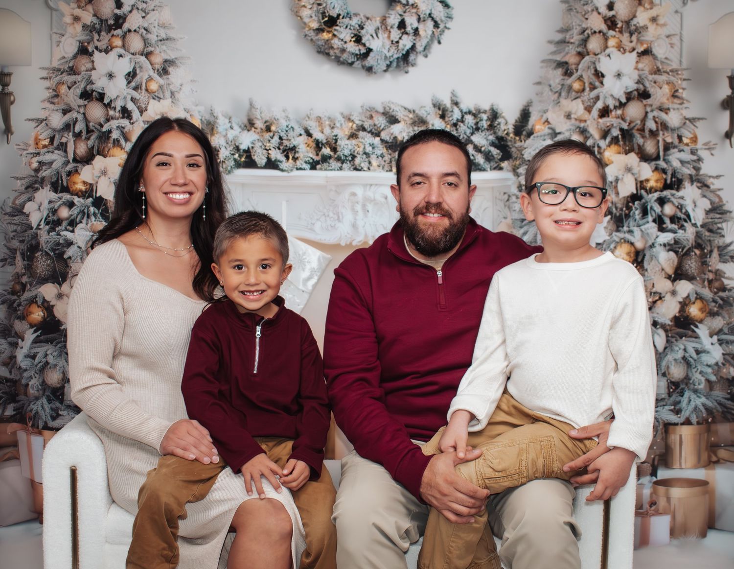 Family of four in neutral tones sitting together in front of decorated Christmas trees for holiday portrait.