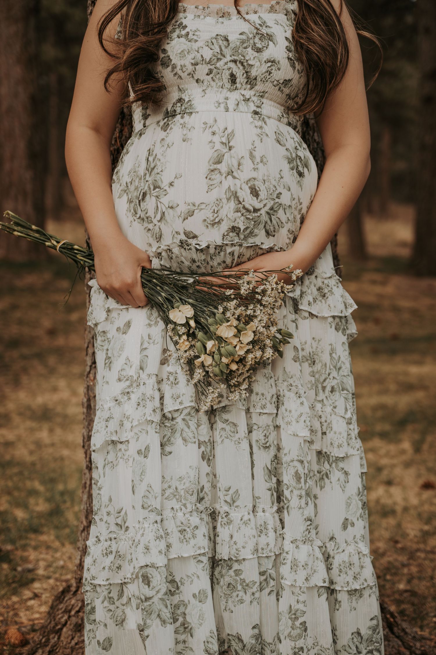 Pregnant woman in white floral tiered maxi dress holding wildflower bouquet outdoors in forest setting.