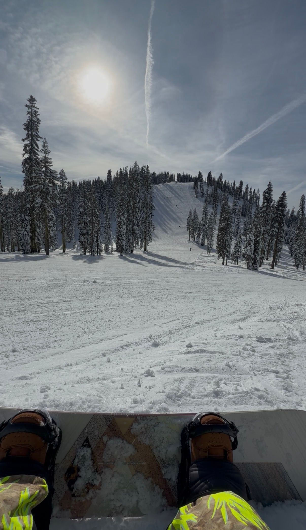 A snowy ski slope with evergreen trees lining the trail under a bright winter sky with sun halo effect.