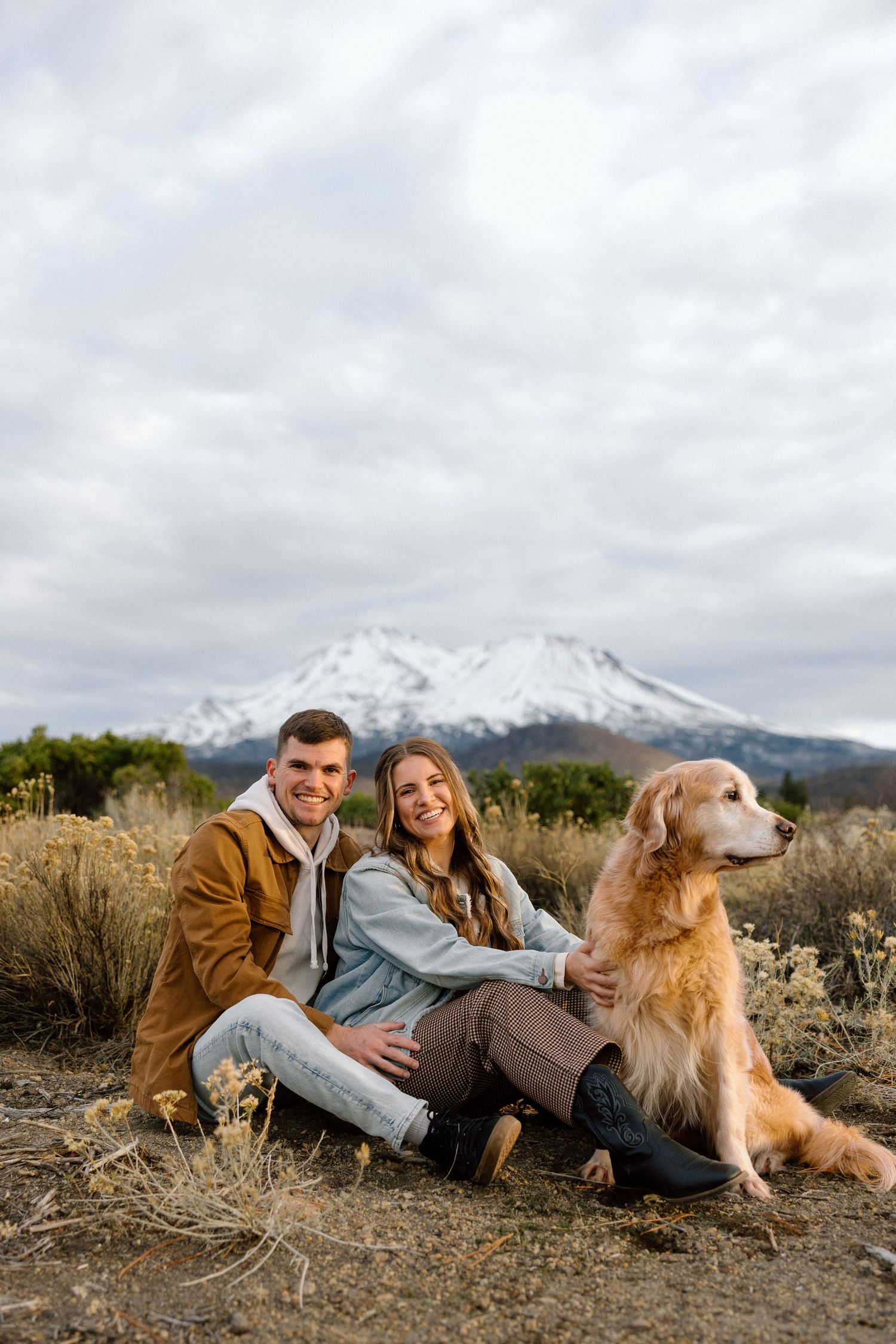 A couple lounges on the ground with their golden retriever dog against a scenic mountain backdrop on a cloudy day.